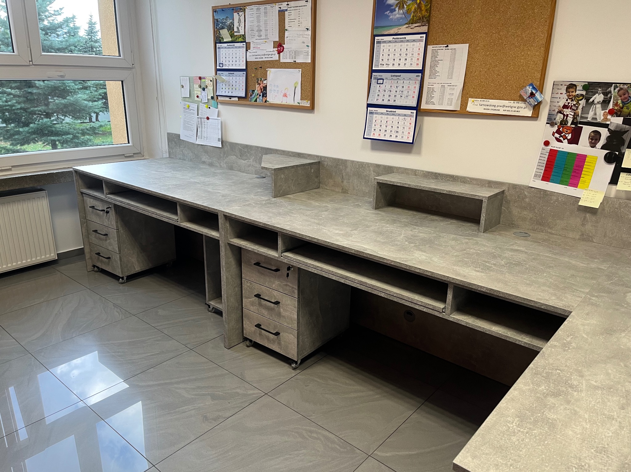 Długi, szary blat biurkowy z szufladami i półkami, zamontowany w biurze. Widoczne kalendarze i tablice korkowe na ścianie. Podłoga wyłożona szarymi płytkami.