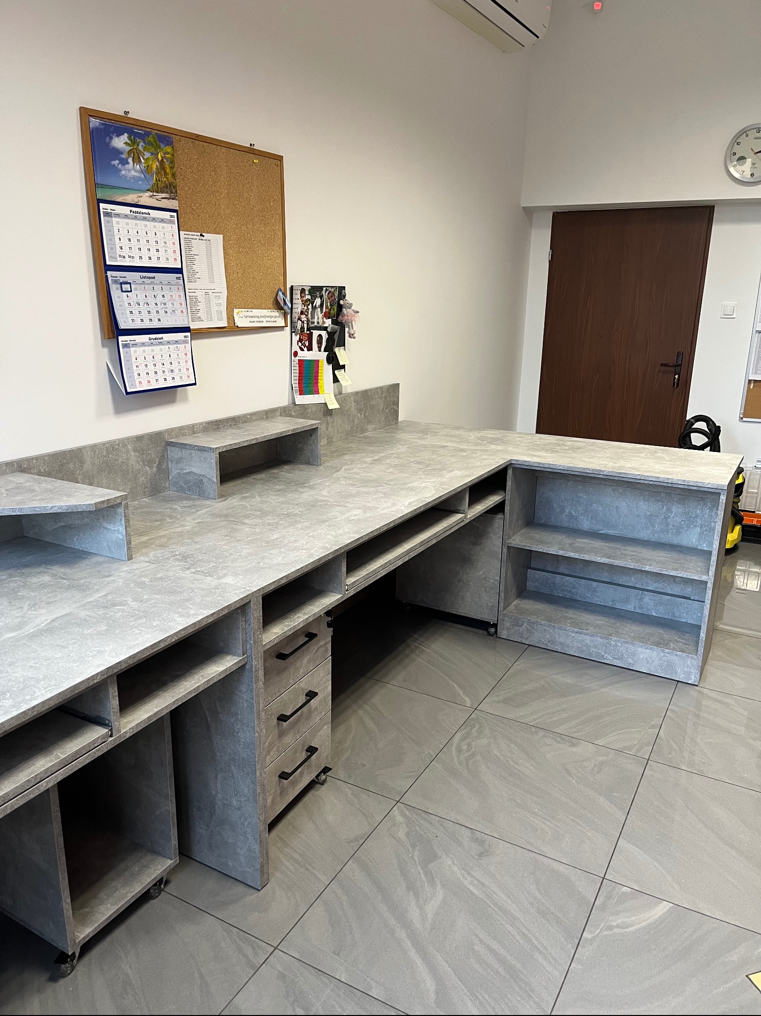 Szary, narożny blat z szafkami i szufladami na kółkach, z kamiennym wzorem, w jasnym pomieszczeniu z płytkami podłogowymi i kalendarzem na ścianie.