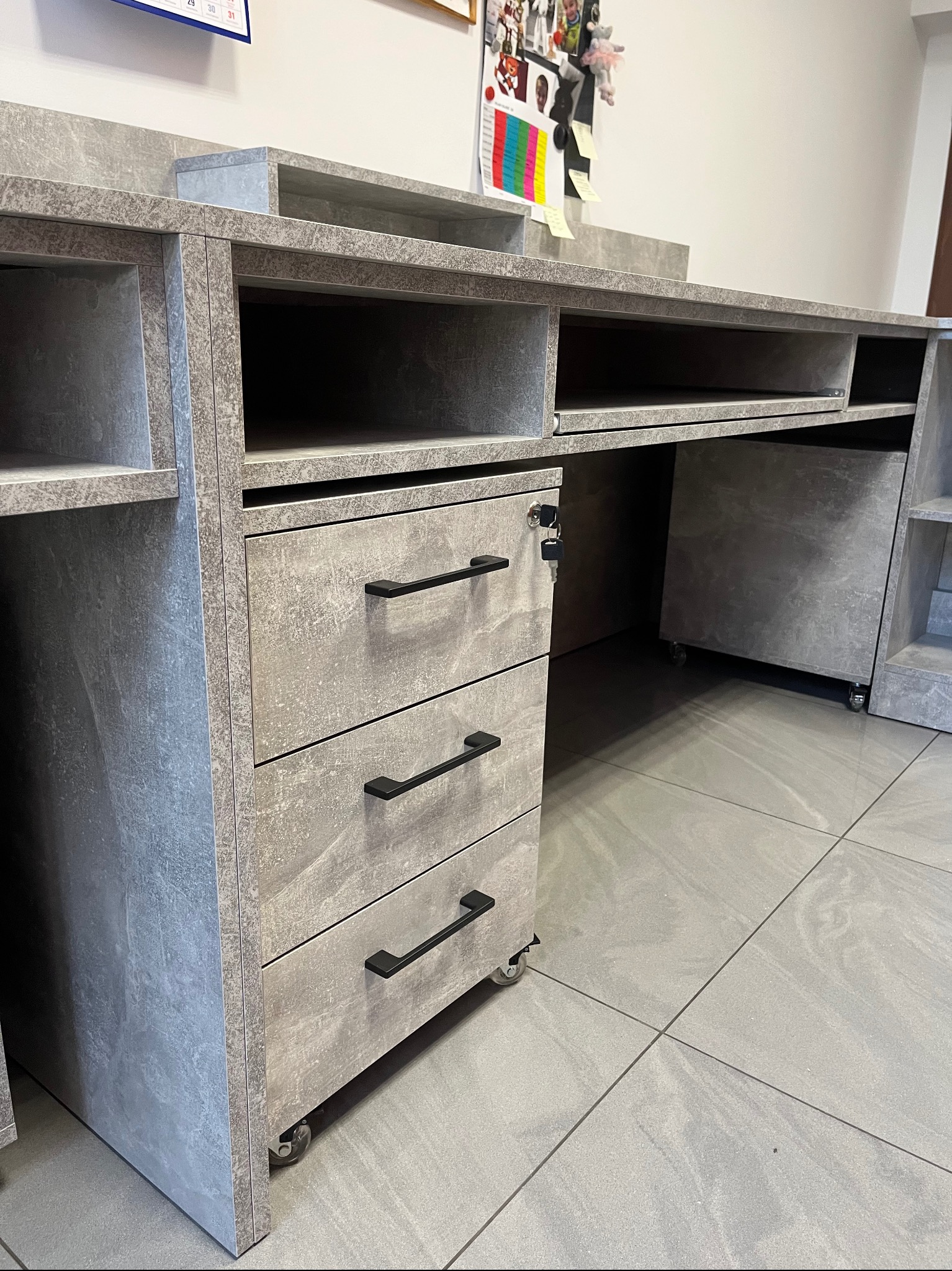 Szara, nowoczesna szafka biurowa z trzema szufladami na kółkach, z czarnymi uchwytami, widoczna pod kątem, na tle szarej podłogi. Klucz w zamku szuflady.