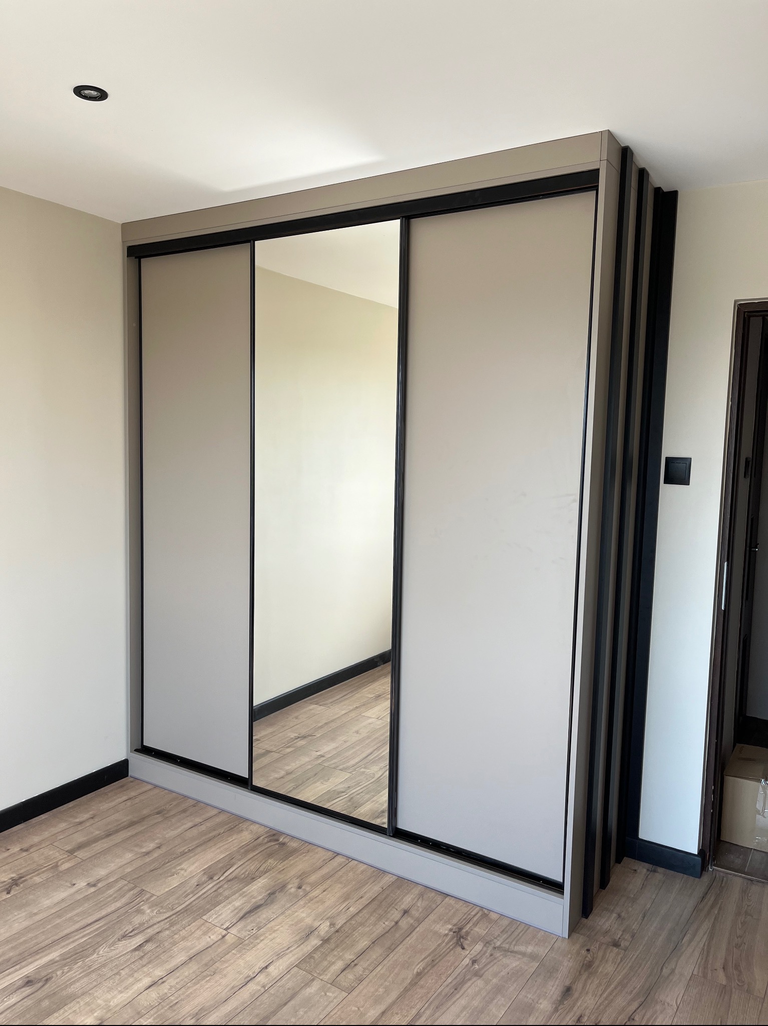 Jasna szafa wnękowa z przesuwnymi drzwiami i lustrem, wykończona czarnymi detalami, na tle drewnianej podłogi. Widoczne pionowe panele dekoracyjne po prawej stronie.