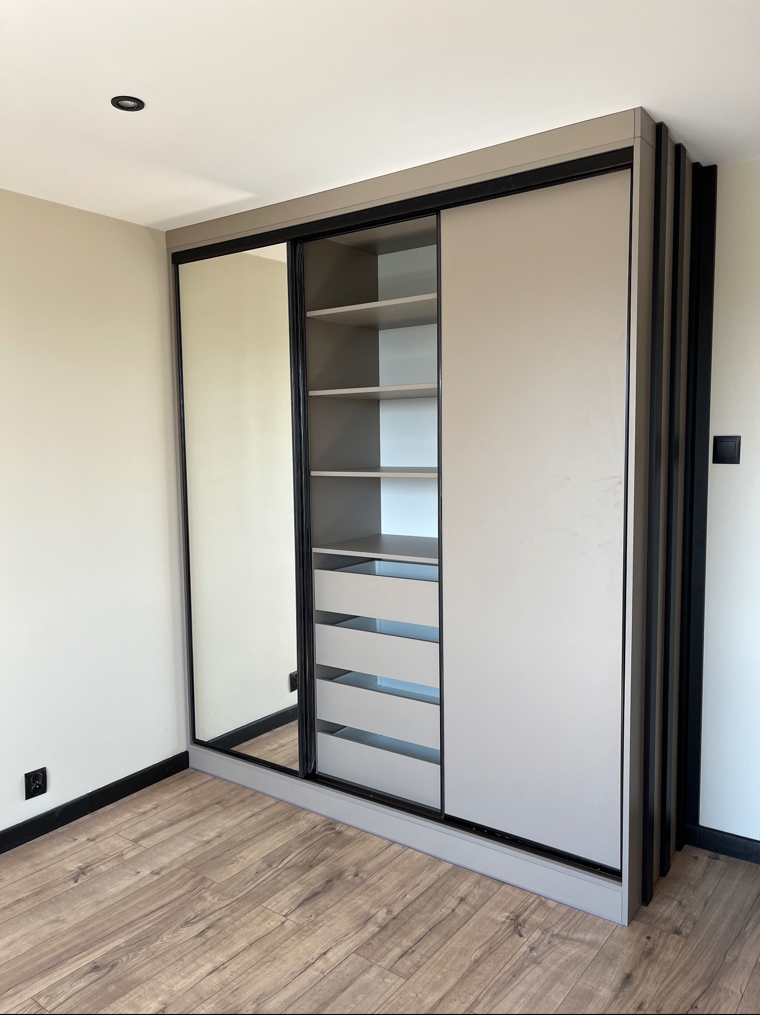 Beżowa szafa wnękowa z przesuwnymi drzwiami i lustrem, czarne akcenty, drewniana podłoga. Nowoczesny design, minimalistyczne wnętrze.