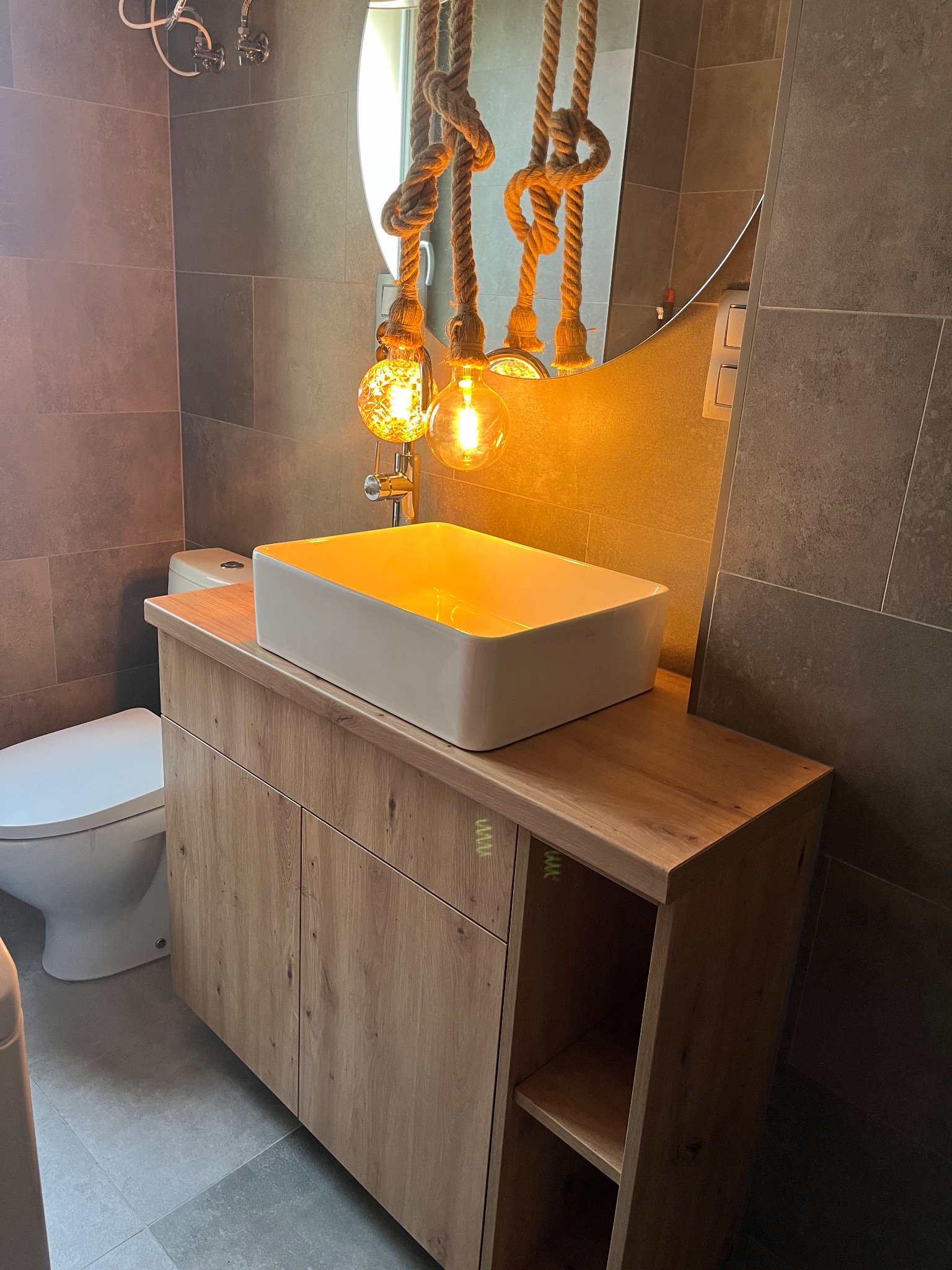 Nowoczesna łazienka z drewnianą szafką pod umywalkę, białą umywalką nablatową i dekoracyjnym oświetleniem na sznurach. Szare płytki na ścianach i podłodze.