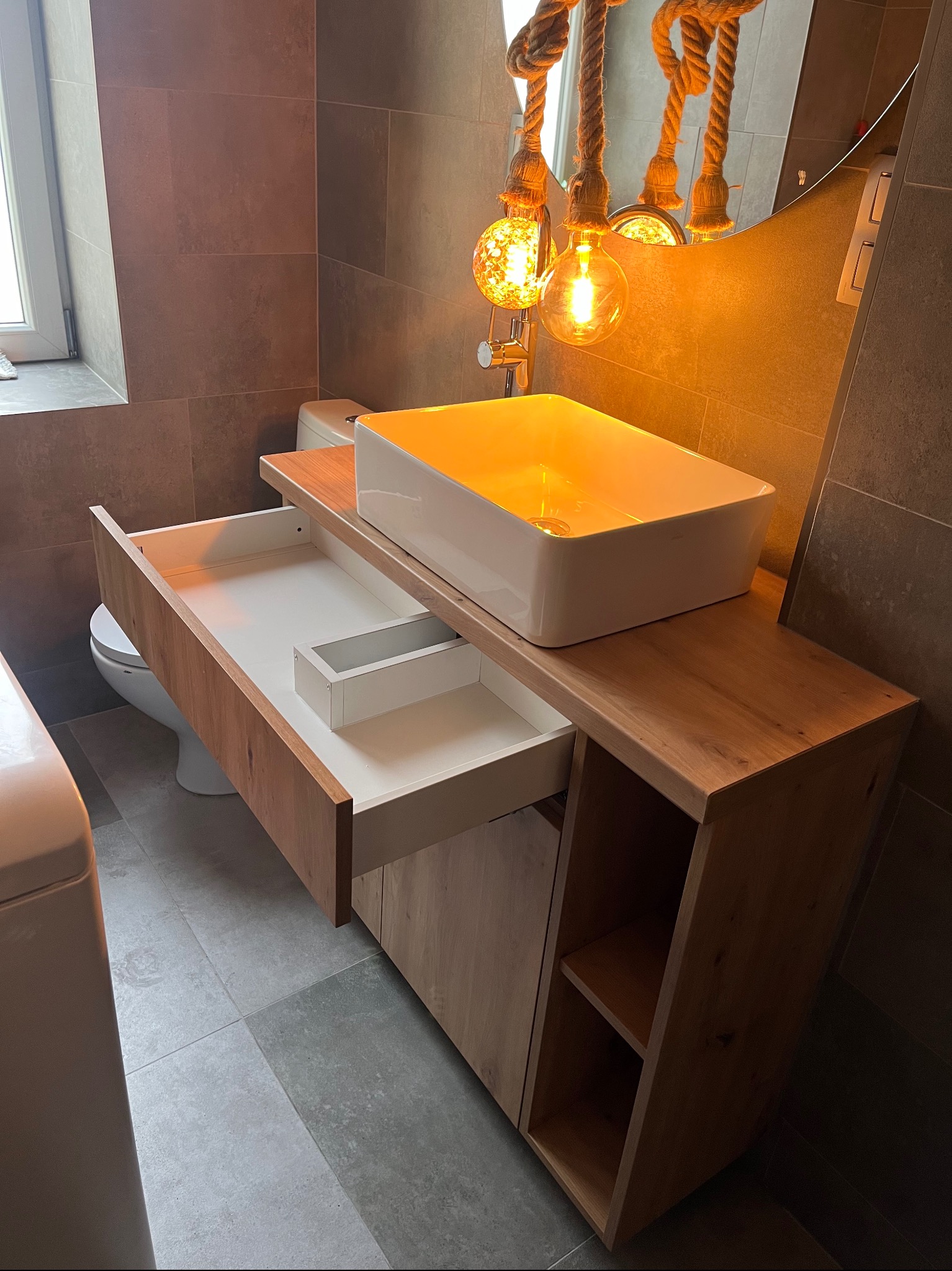 Nowoczesna szafka łazienkowa z blatem drewnianym i białą umywalką nablatową. Otwarta szuflada z białym wnętrzem. Dekoracyjne oświetlenie z żarówkami Edison.