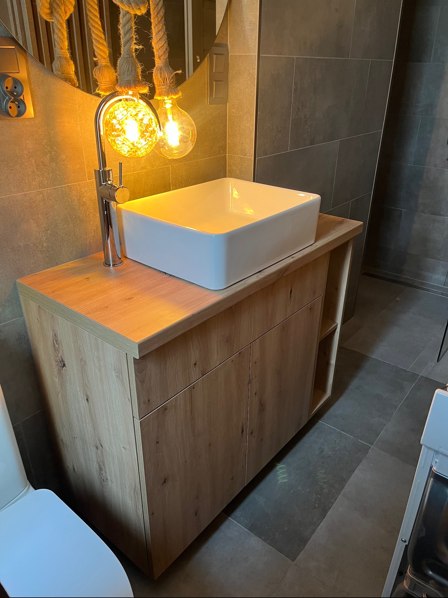 Nowoczesna łazienka: szafka z blatem z jasnego drewna pod umywalką nablatową, oświetlona dekoracyjnymi żarówkami na tle szarych płytek.