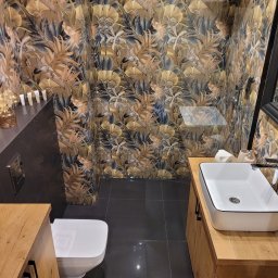 RENOVATION Piotr Kwiatkowski - Nowoczesna łazienka z toaletą, umywalką nablatową na drewnianej szafce i ścianami wyłożonymi płytkami z motywem liści w odcieniach brązu i szarości.
