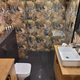 Nowoczesna łazienka z toaletą, umywalką nablatową na drewnianej szafce i ścianami wyłożonymi płytkami z motywem liści w odcieniach brązu i szarości.