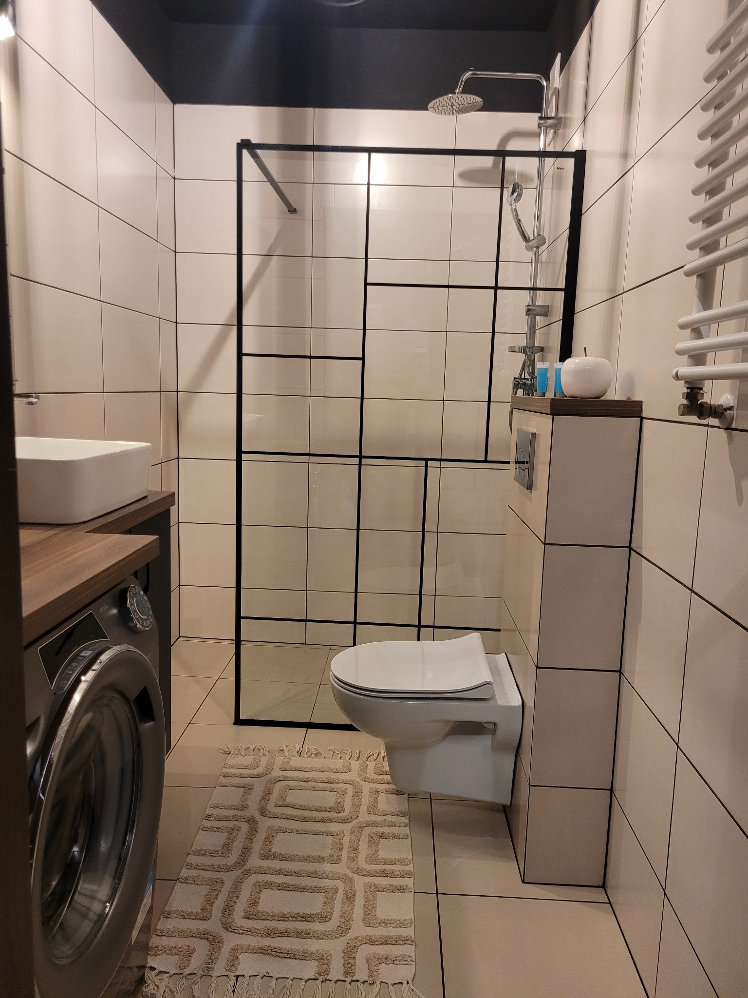 Nowoczesna łazienka z kabiną prysznicową typu walk-in z czarnymi profilami, umywalką nablatową na drewnianym blacie, pralką, podwieszaną toaletą i beżowymi płytkami kwadratowymi.