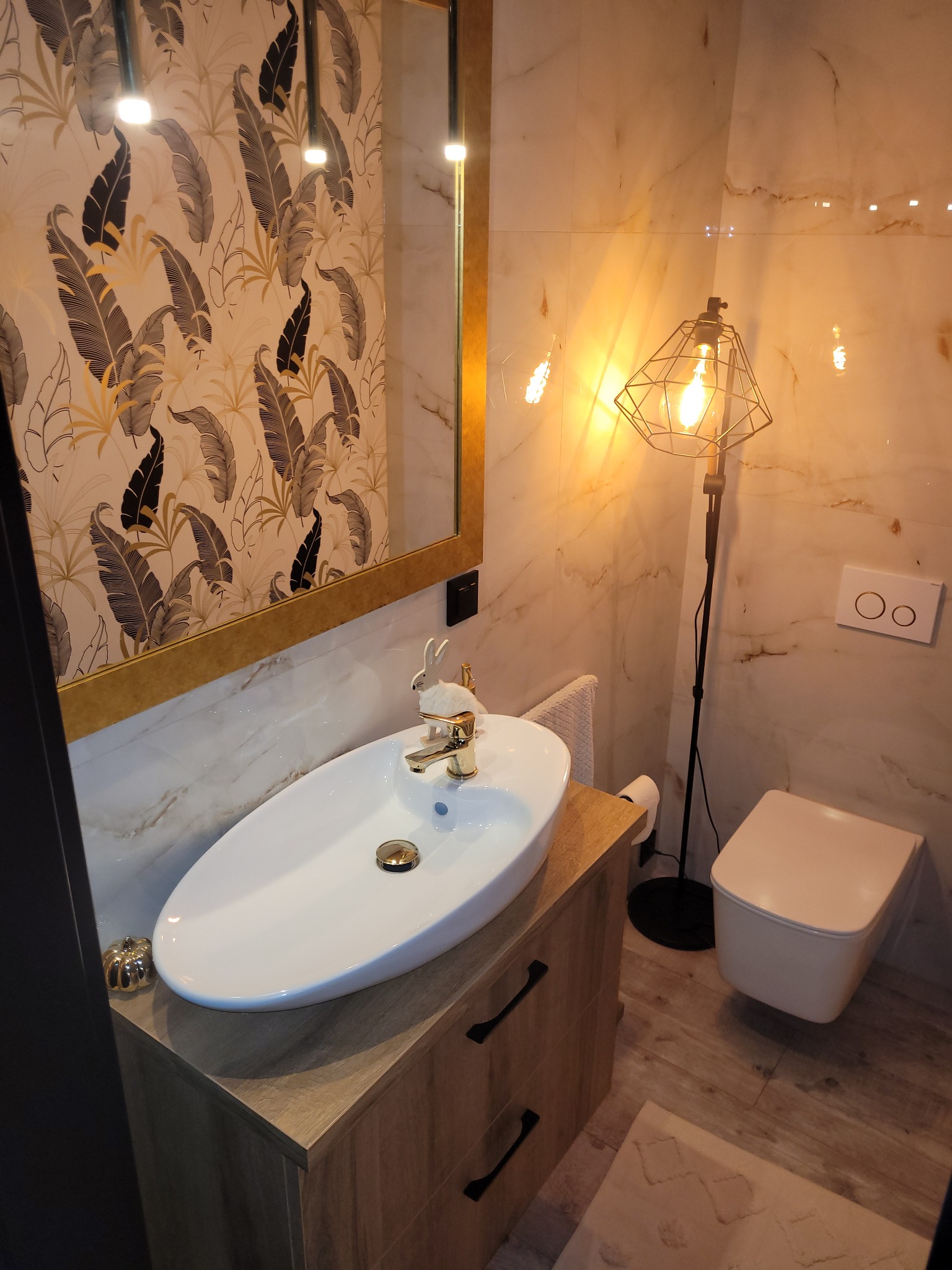 Nowoczesna łazienka z umywalką nablatową, złotą armaturą i dekoracyjnym oświetleniem, marmurowe płytki na ścianach, ozdobny królik na umywalce.