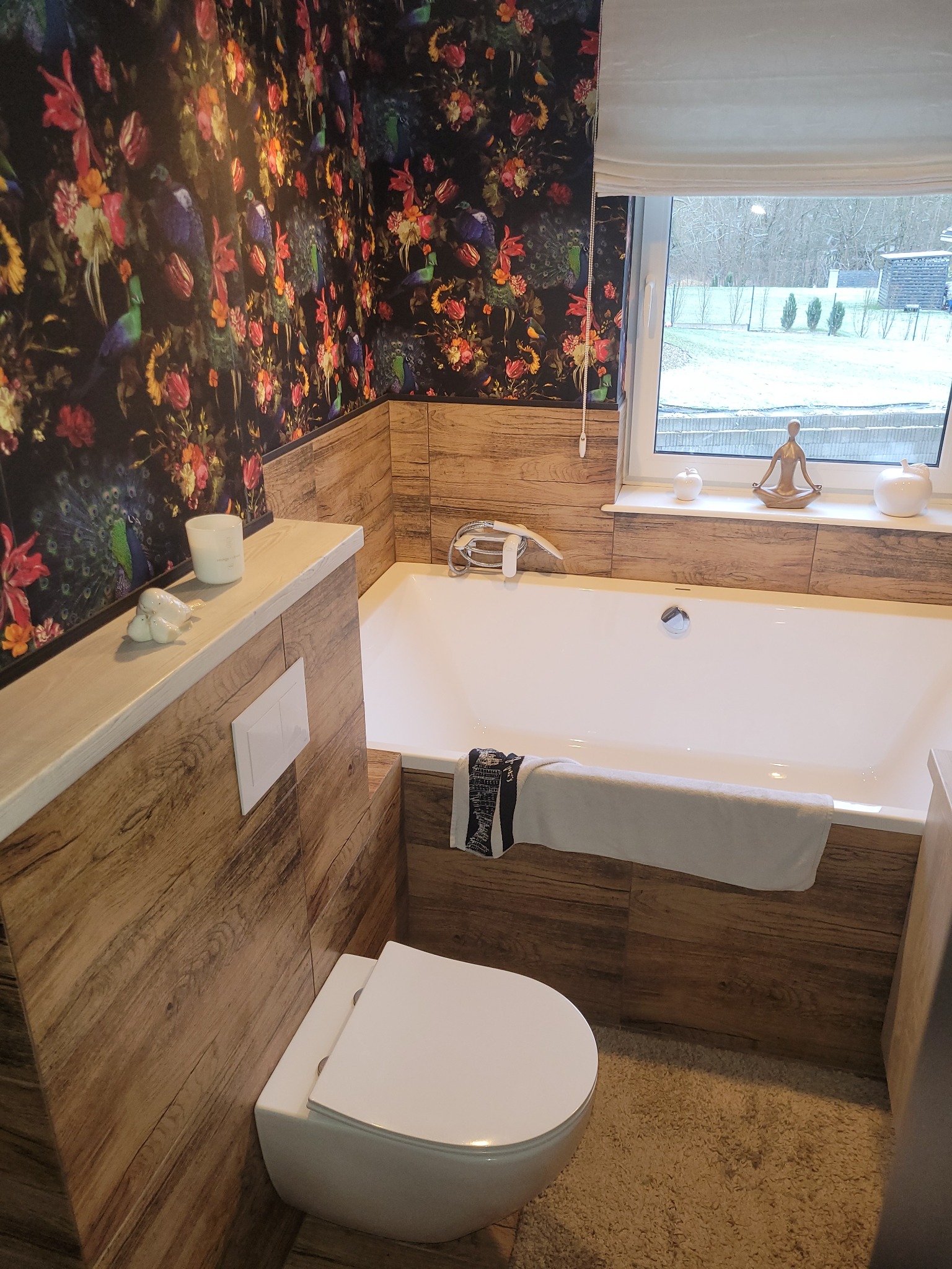 Nowoczesna łazienka z wanną obudowaną płytkami imitującymi drewno, toaleta podwieszana, ściana z wzorzystą tapetą w kwiaty i pawie, okno z dekoracjami, dywanik z długim włosiem.