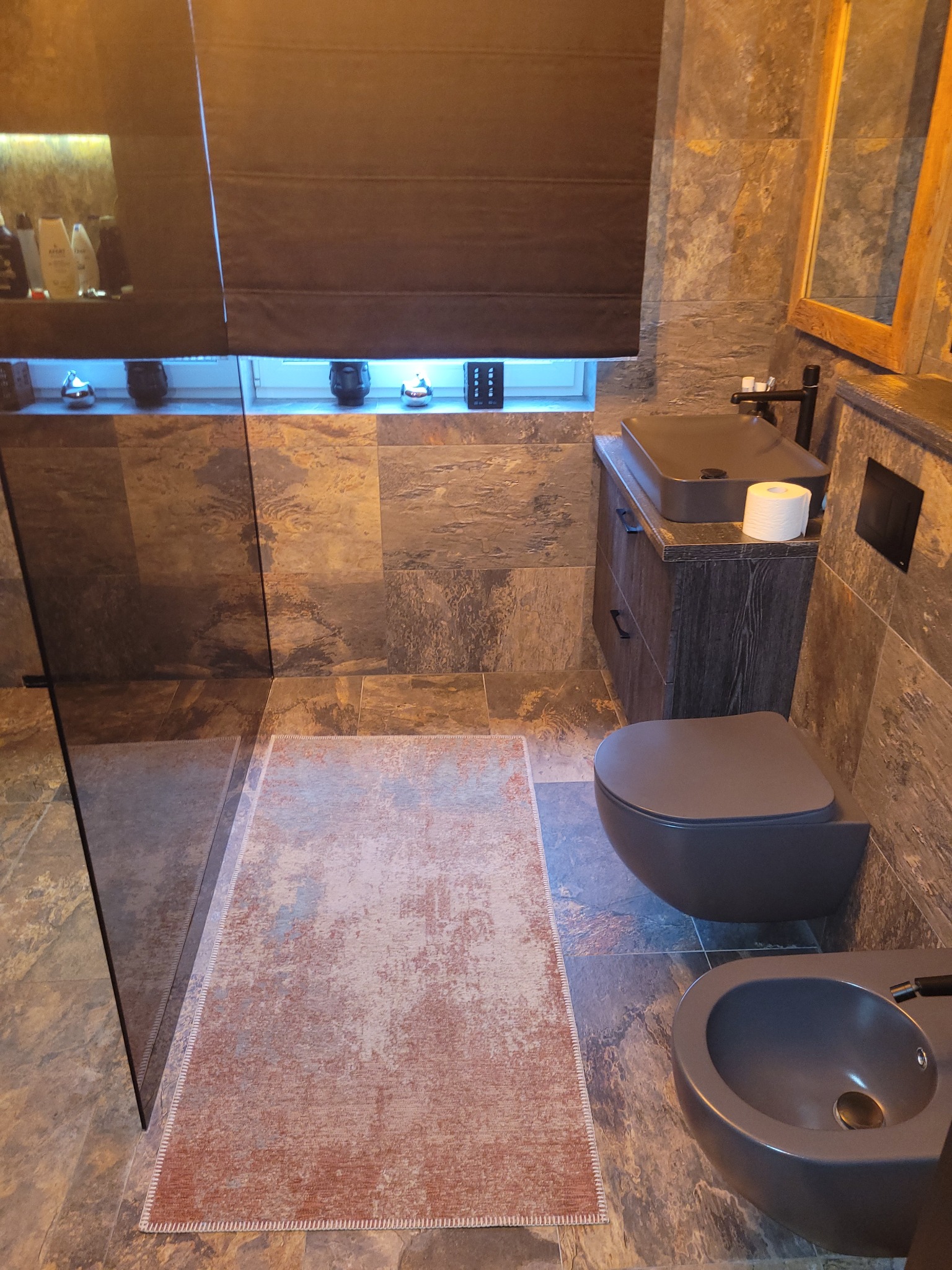 Nowoczesna łazienka w odcieniach szarości z deszczownicą, wiszącą toaletą i bidetem, wykończona płytkami imitującymi kamień, oświetlona ciepłym światłem, z dywanikiem w kolorze terakoty.