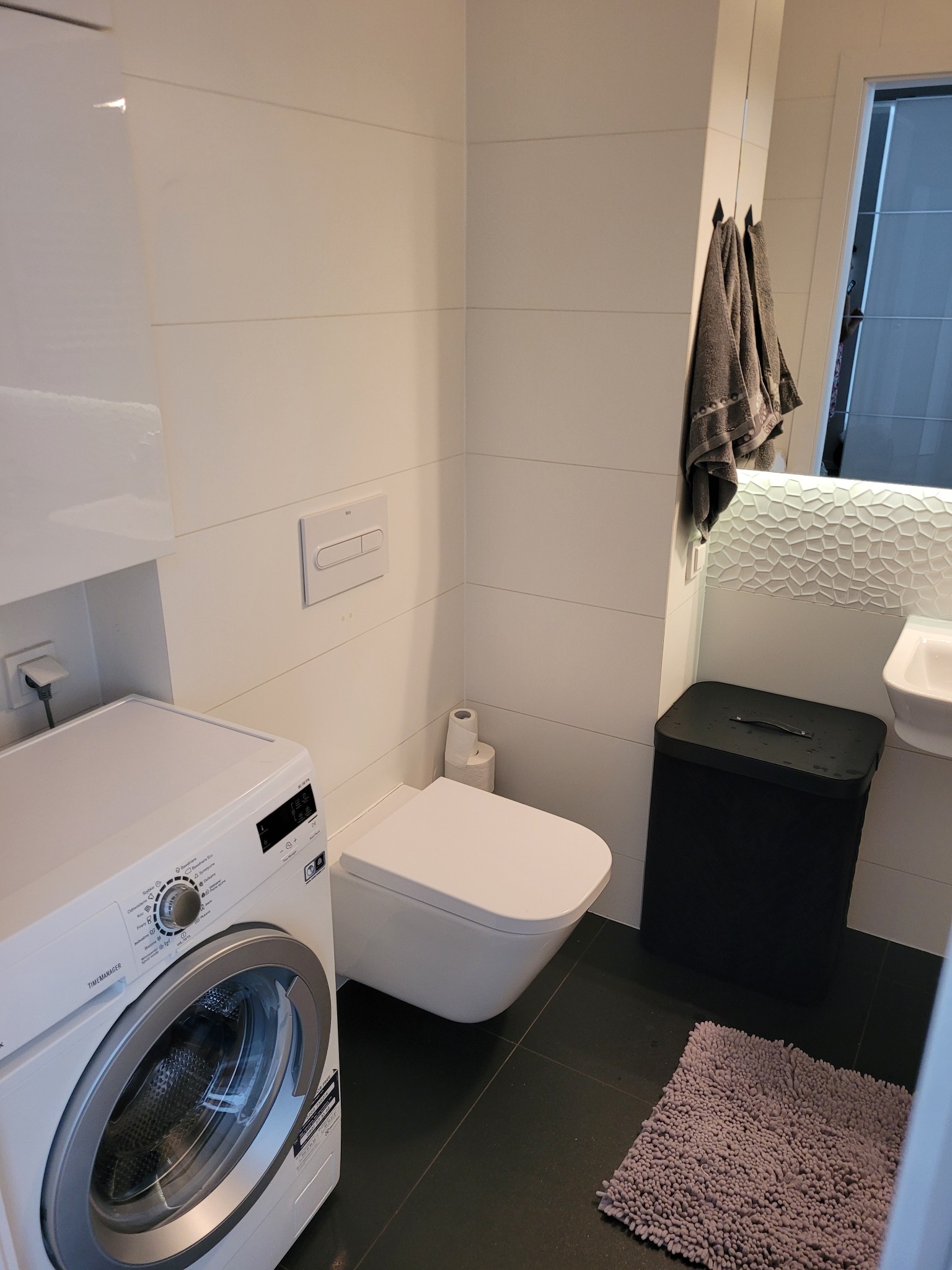 Nowoczesna łazienka z białą pralką, podwieszaną toaletą, szarą wycieraczką i lustrem z dekoracyjnym oświetleniem.