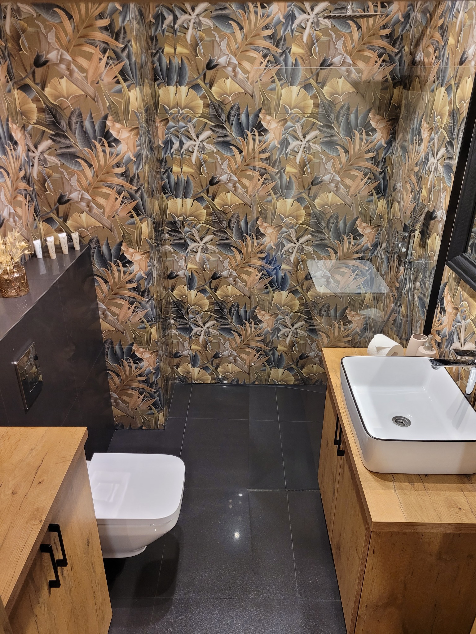 Nowoczesna łazienka z toaletą, umywalką nablatową na drewnianej szafce i ścianami wyłożonymi płytkami z motywem liści w odcieniach brązu i szarości.