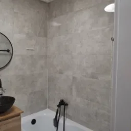 Szara łazienka z wanną, umywalką na drewnianej szafce i okrągłym lustrem. Płytki imitujące kamień na ścianach. Czarna armatura.