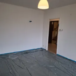 Pokój po malowaniu na biało, zabezpieczony folią malarską i taśmą. Widoczna lampa, gniazdka i fragment schodów w tle. Świeżo pomalowane ściany.