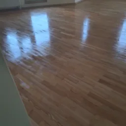 Parkiet po renowacji, lakierowany na wysoki połysk, odbijający światło z okna. Widoczny grzejnik i fragment ściany w jasnym kolorze.