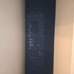 Niebieska, błyszcząca mozaika ułożona w wąskiej wnęce łazienki, sąsiadująca z jasnymi płytkami ściennymi. Widoczne fugi i odbicia światła na powierzchni płytek.