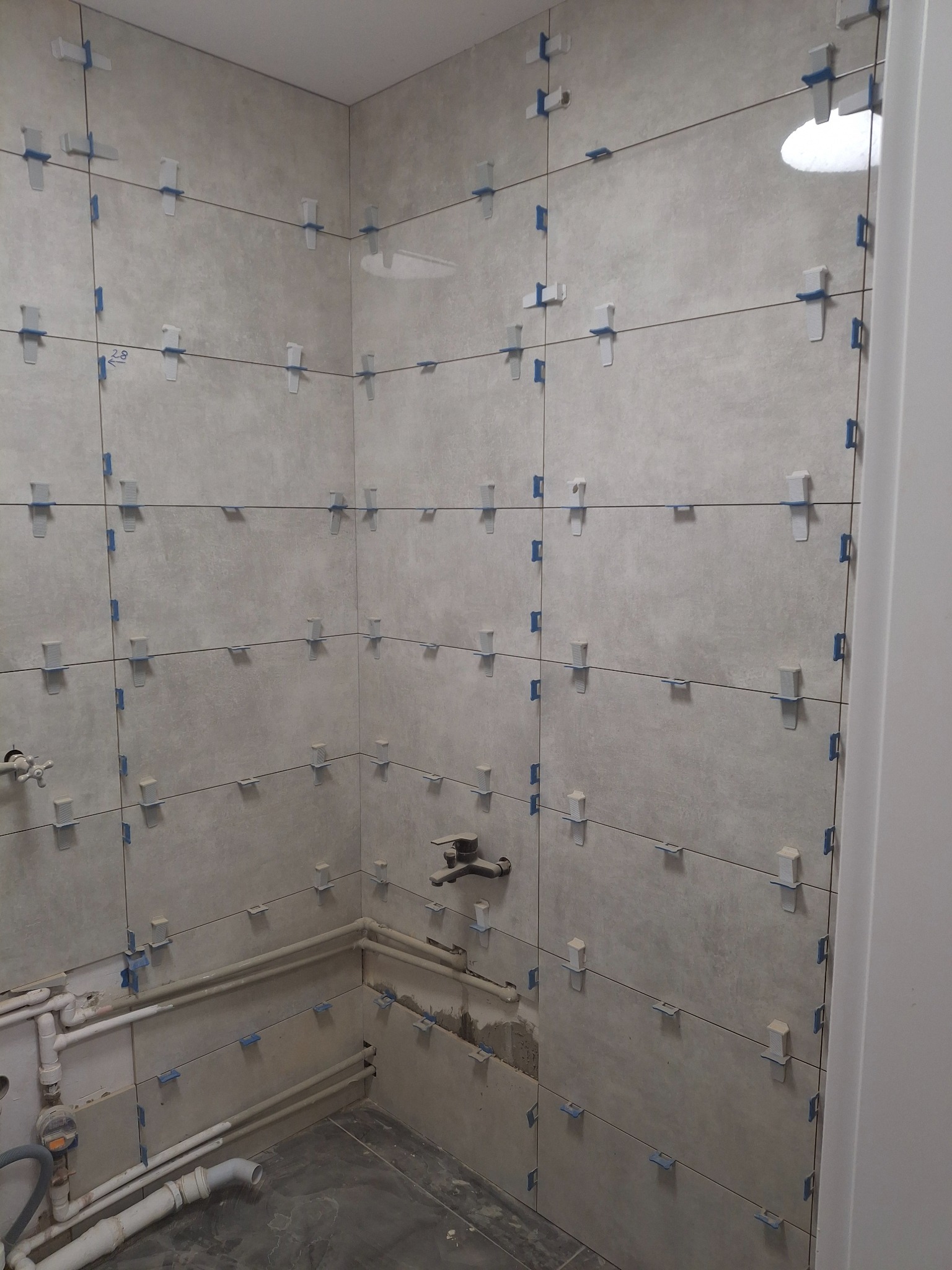 Szare płytki ścienne w trakcie montażu w rogu łazienki, z widocznymi krzyżykami dystansowymi i fragmentem instalacji hydraulicznej w dolnej części.