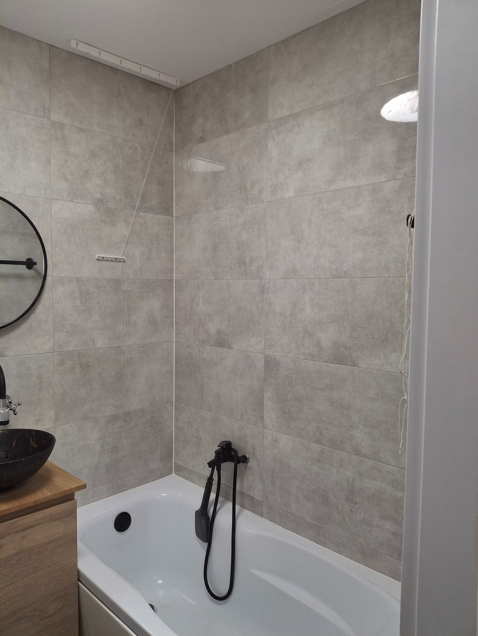 Szara łazienka z wanną, umywalką na drewnianej szafce i okrągłym lustrem. Płytki imitujące kamień na ścianach. Czarna armatura.