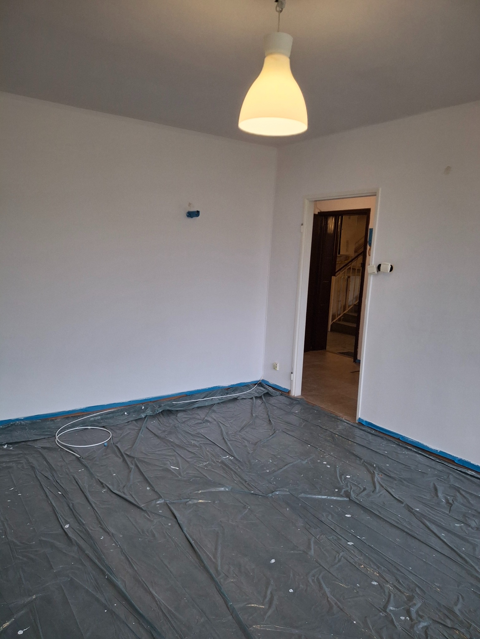Pokój po malowaniu na biało, zabezpieczony folią malarską i taśmą. Widoczna lampa, gniazdka i fragment schodów w tle. Świeżo pomalowane ściany.
