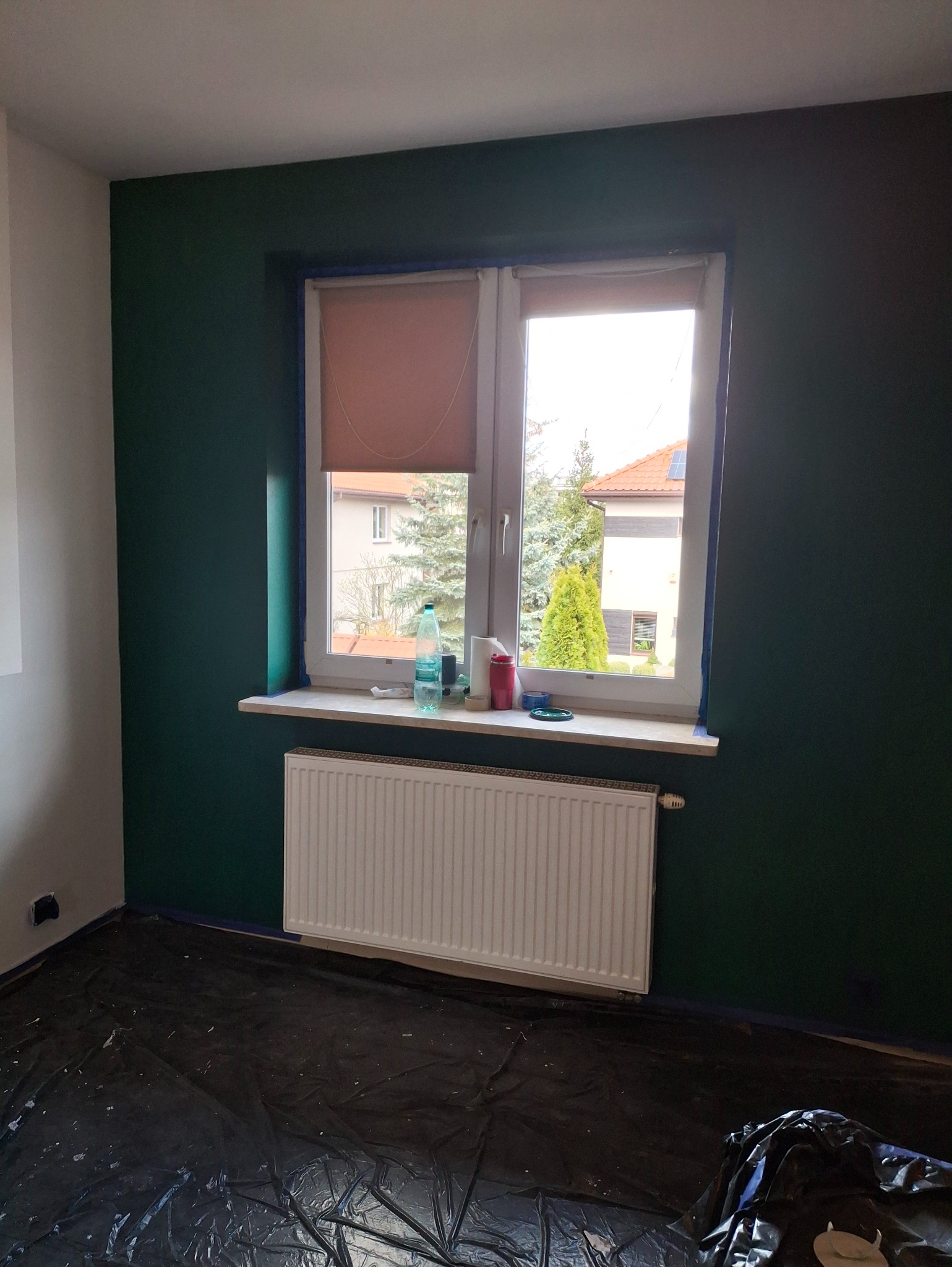 Pokój po malowaniu ścian na ciemnozielono, okno z roletą, grzejnik, zabezpieczona folią podłoga. Widoczne akcesoria malarskie na parapecie.