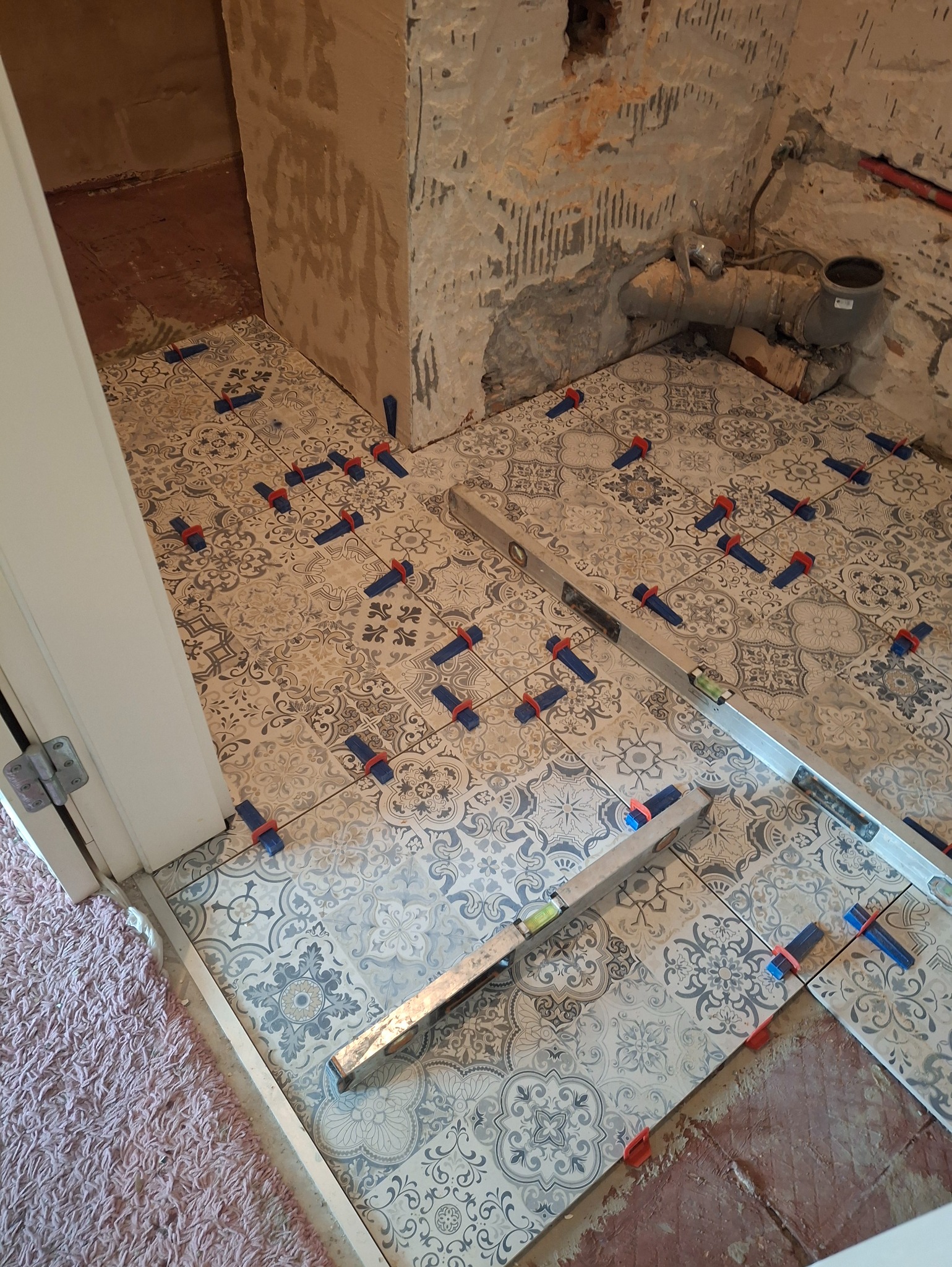 Układanie wzorzystych płytek podłogowych w łazience, z użyciem poziomicy i klipsów do poziomowania. Widoczne surowe ściany i instalacja kanalizacyjna.