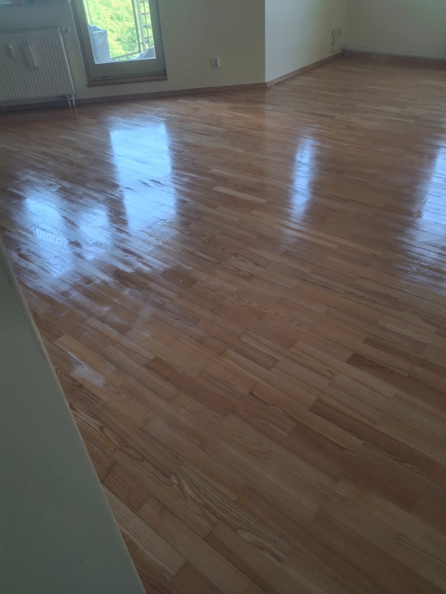 Parkiet po renowacji, lakierowany na wysoki połysk, odbijający światło z okna. Widoczny grzejnik i fragment ściany w jasnym kolorze.