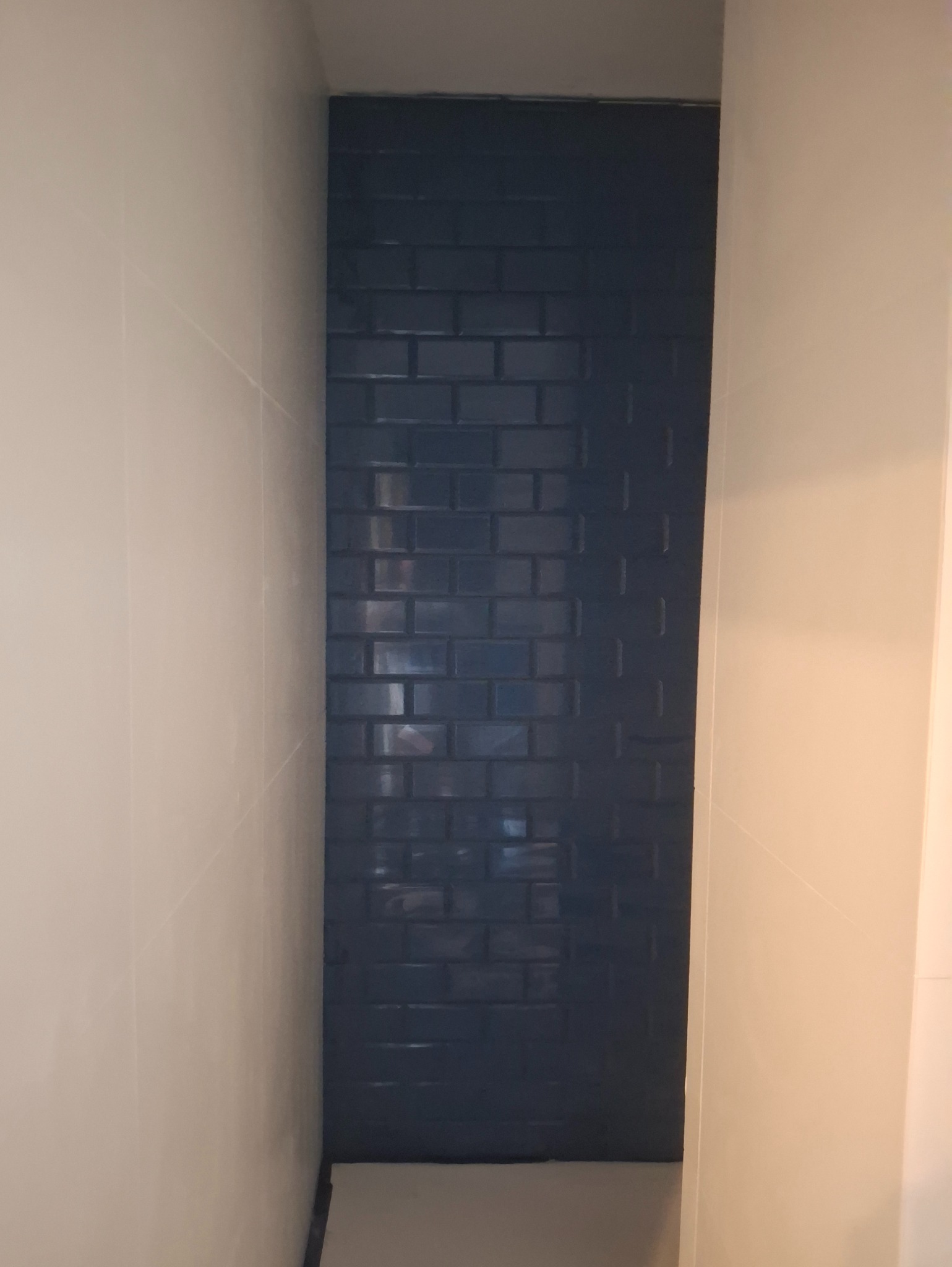 Niebieska, błyszcząca mozaika ułożona w wąskiej wnęce łazienki, sąsiadująca z jasnymi płytkami ściennymi. Widoczne fugi i odbicia światła na powierzchni płytek.