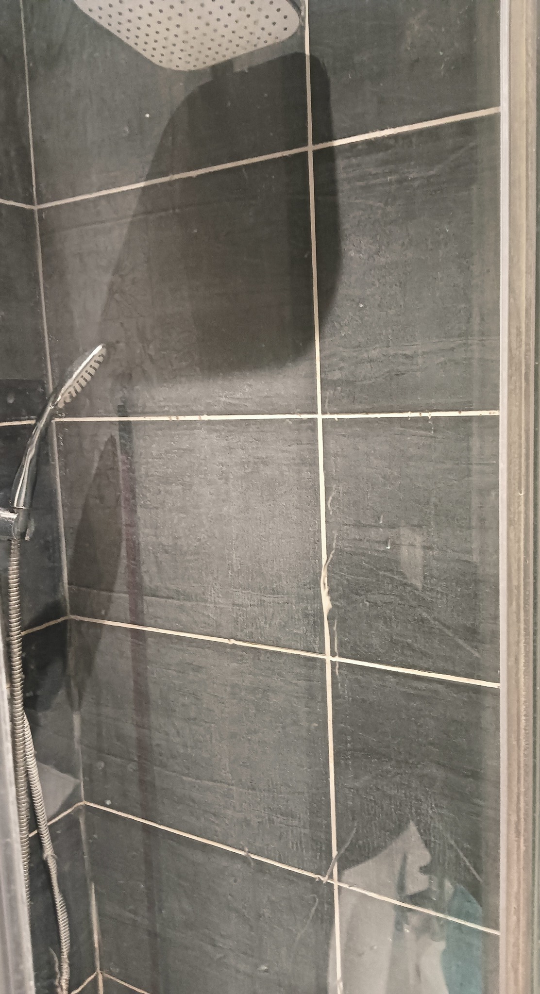 Brudna kabina prysznicowa z ciemnymi płytkami, widoczne osady i zacieki na szkle oraz płytkach, wymagająca czyszczenia.