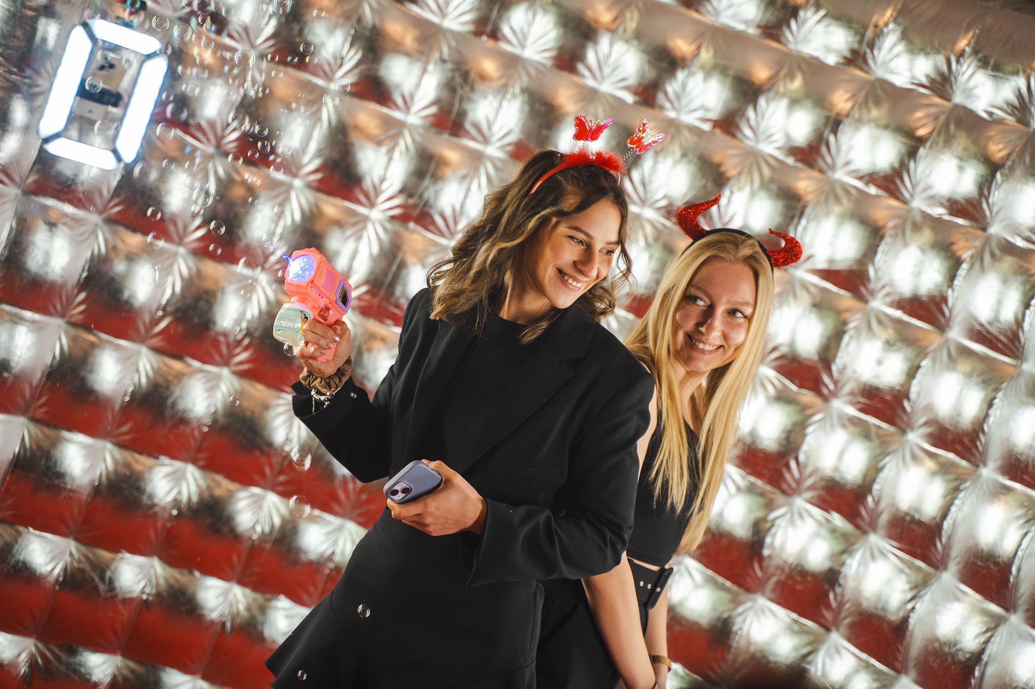 Dwie uśmiechnięte kobiety w kostiumach z akcesoriami (opaski z rogami i motylami) bawią się w fotobudce z srebrnym tłem i puszczają bańki mydlane.