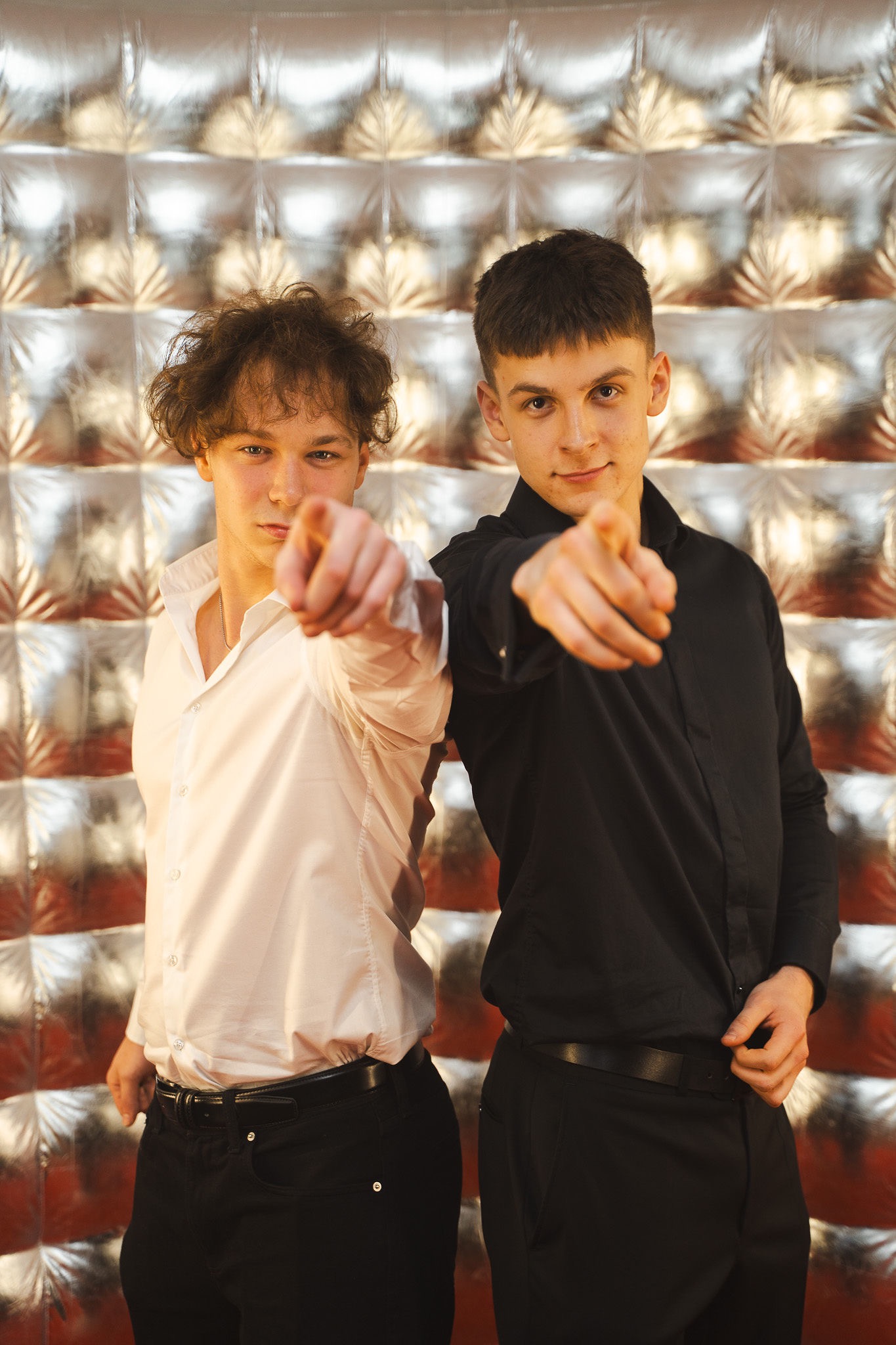 Dwóch młodych mężczyzn, jeden w białej, drugi w czarnej koszuli, wskazują palcami w stronę widza na tle srebrnej, błyszczącej ściany. Fotografia budki.