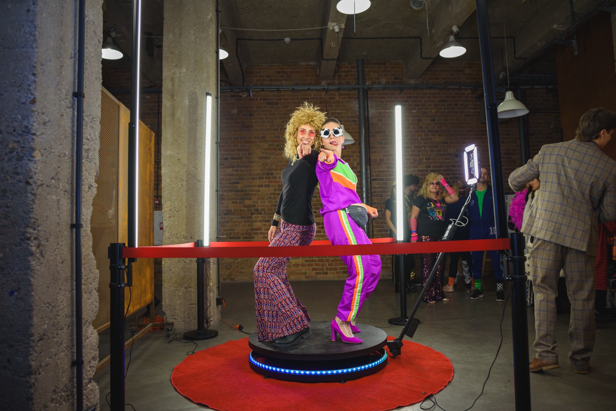 Dwie kobiety w kostiumach retro pozują na obrotowej platformie z oświetleniem LED, otoczone przez inne osoby i elementy scenografii w pomieszczeniu z ceglanymi ścianami.