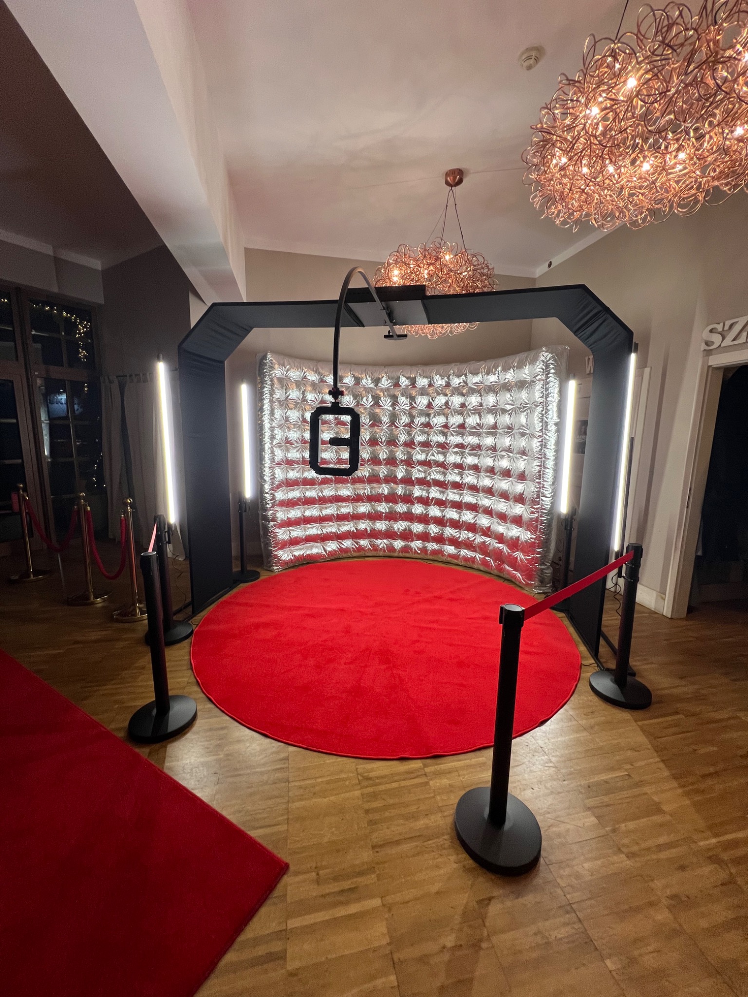 Okrągła fotobudka 360 z czerwonym dywanem, srebrnym tłem i czarną ramą, oświetlona lampami. W tle parkiet i dekoracyjne lampy.