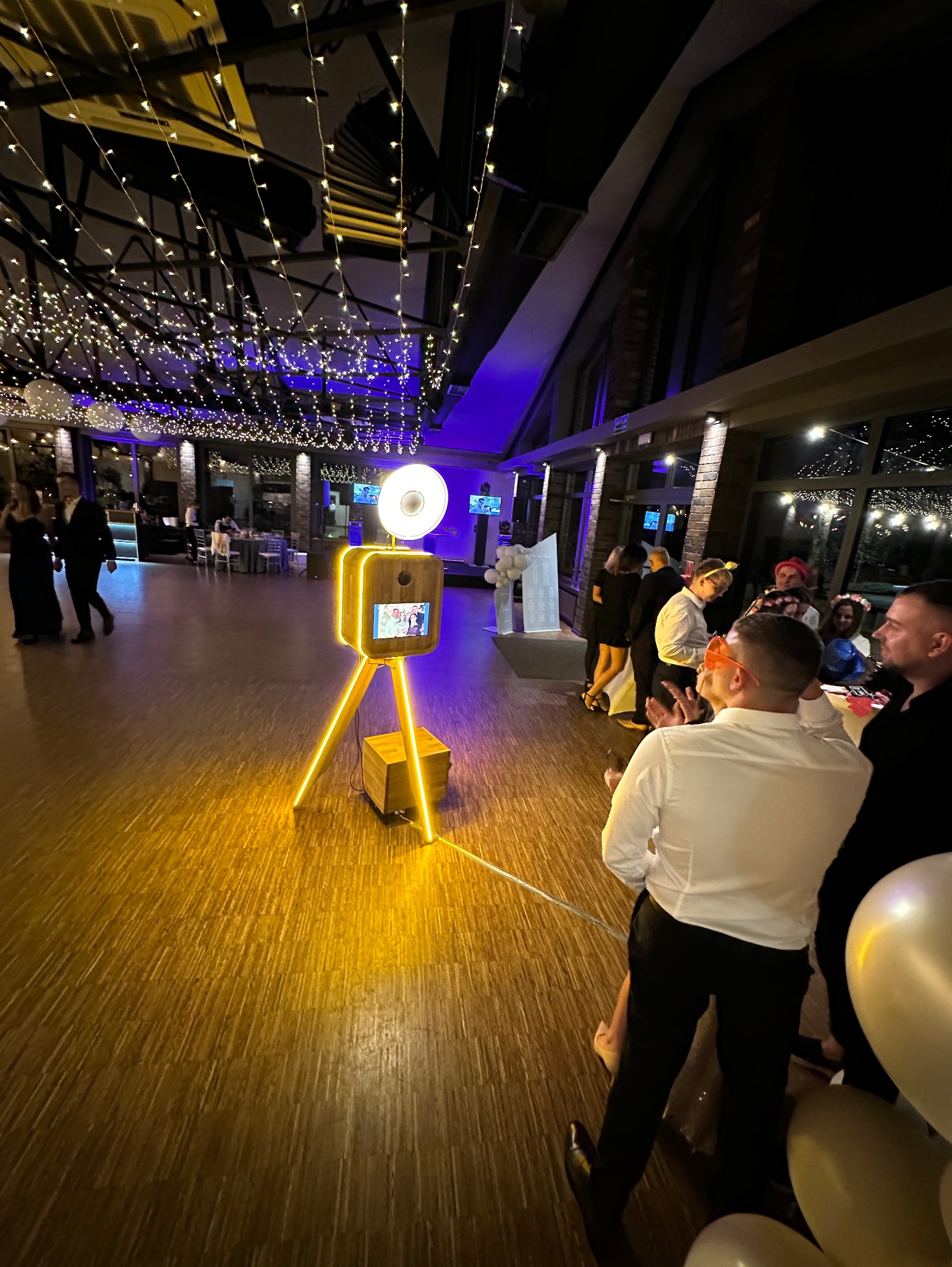 Drewniana fotobudka z neonowym oświetleniem na parkiecie sali balowej, w tle goście weselni i dekoracje świetlne, widok z perspektywy uczestnika zabawy.