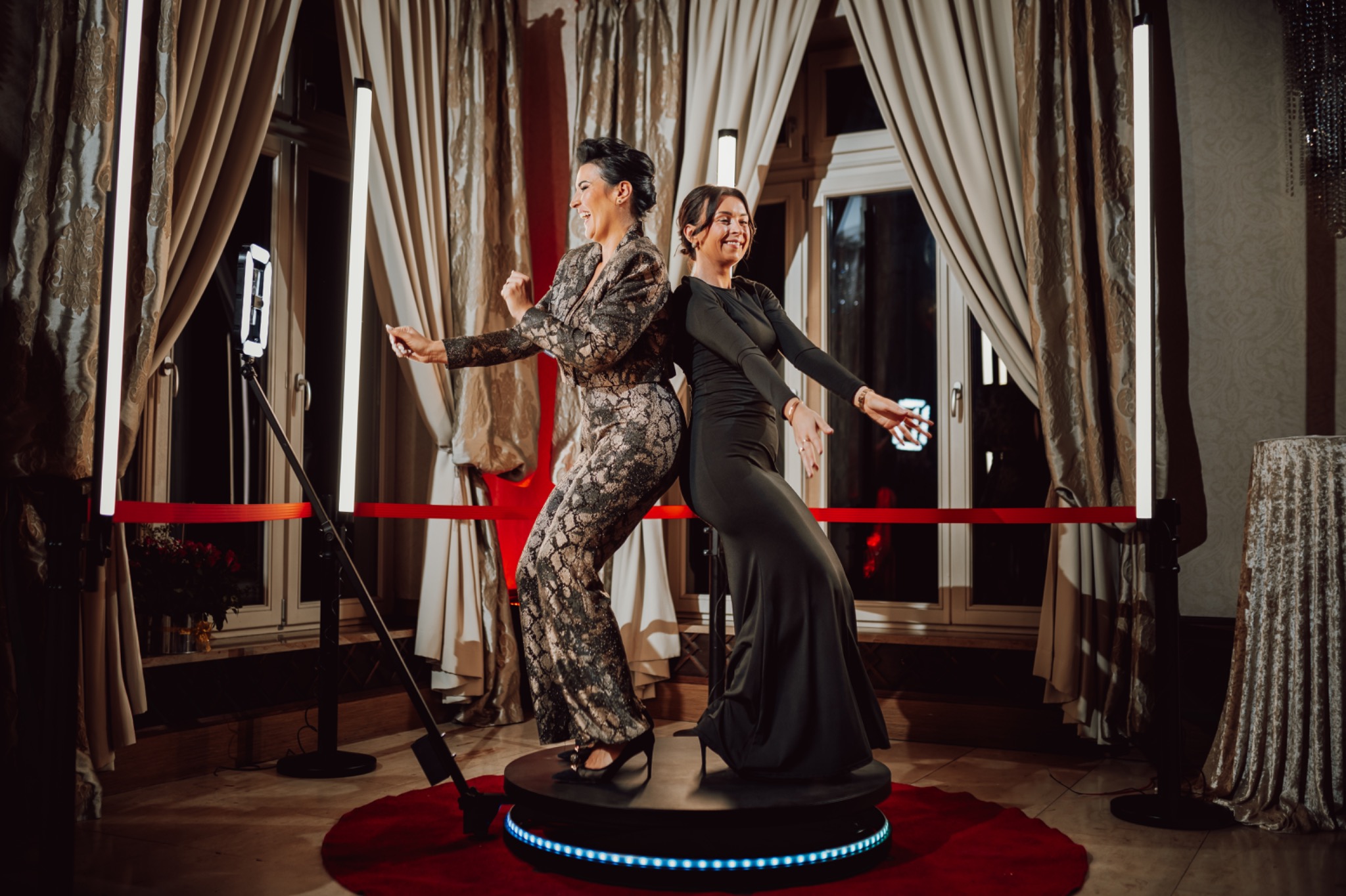 Dwie uśmiechnięte kobiety w eleganckich strojach pozują na obrotowej platformie z niebieskim oświetleniem, otoczonej lampami i czerwonymi barierkami w bogato zdobionym wnętrzu.