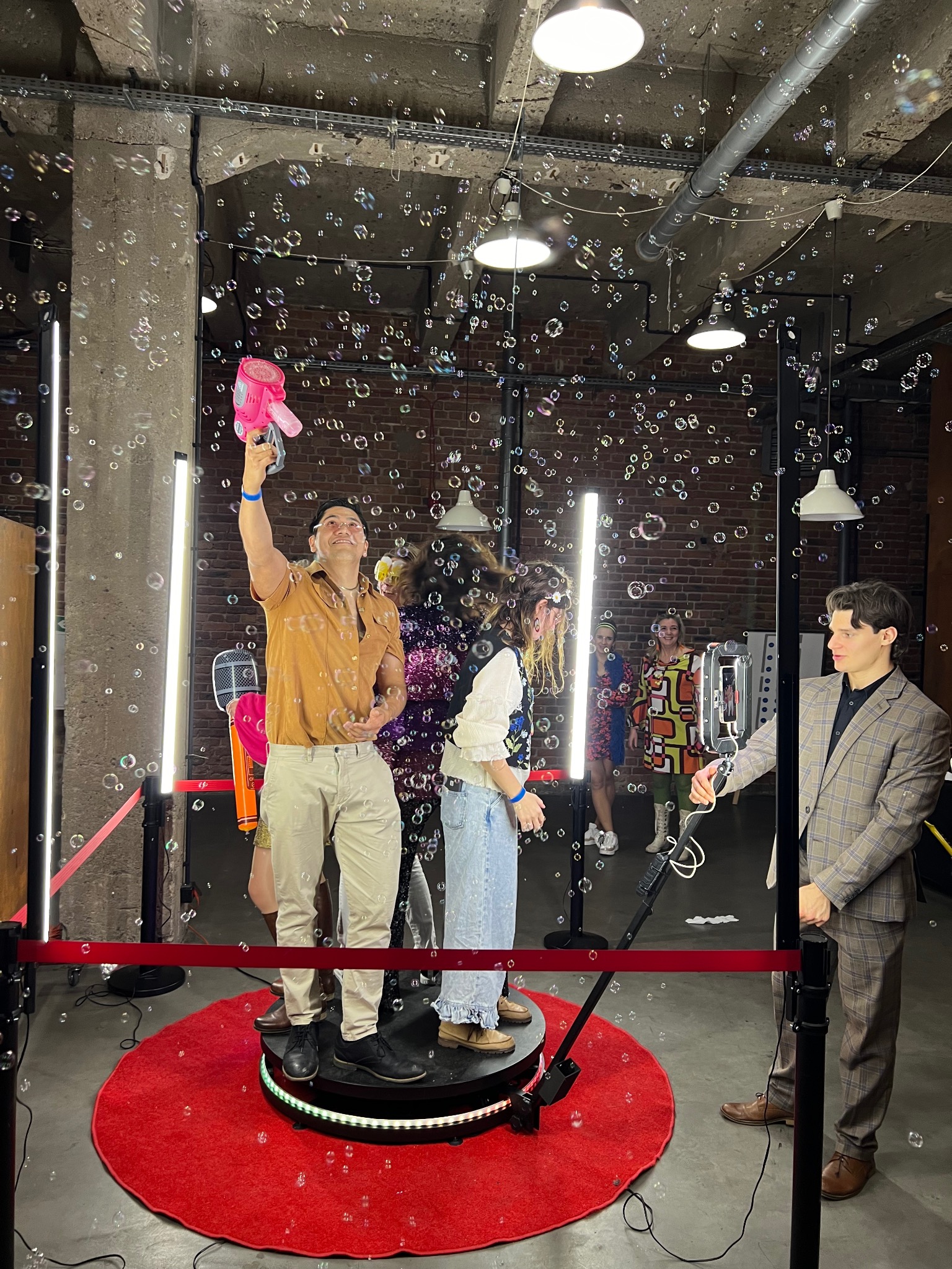 Uczestnicy zabawy w obiektywie obrotowej platformy, otoczeni bańkami mydlanymi i oświetleniem LED, tworzący dynamiczne wideo na tle ceglanej ściany.