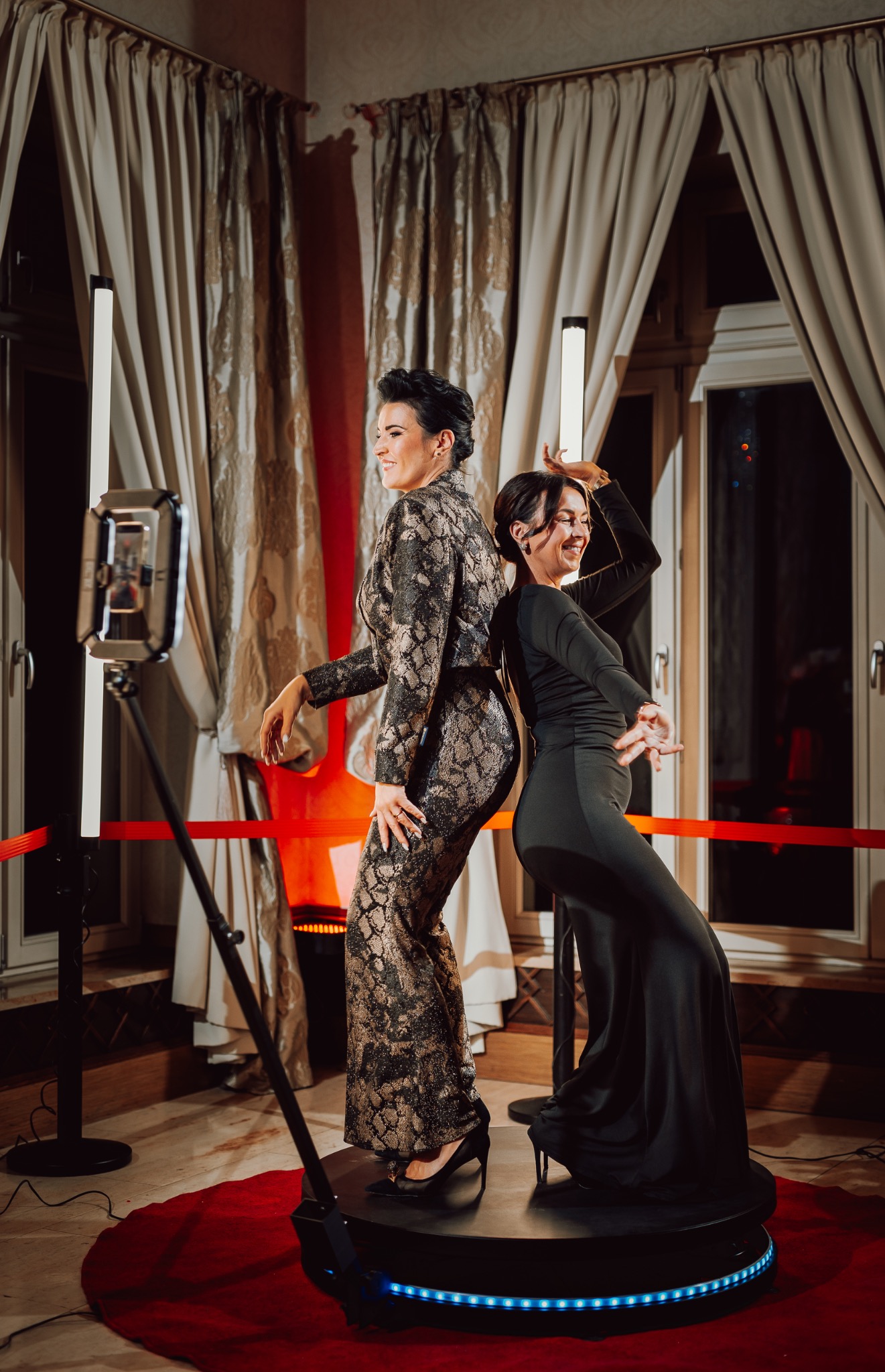 Dwie uśmiechnięte kobiety pozują na platformie 360 fotobudki na tle kurtyn i okna. Jedna w kombinezonie, druga w sukni, obie na obcasach.