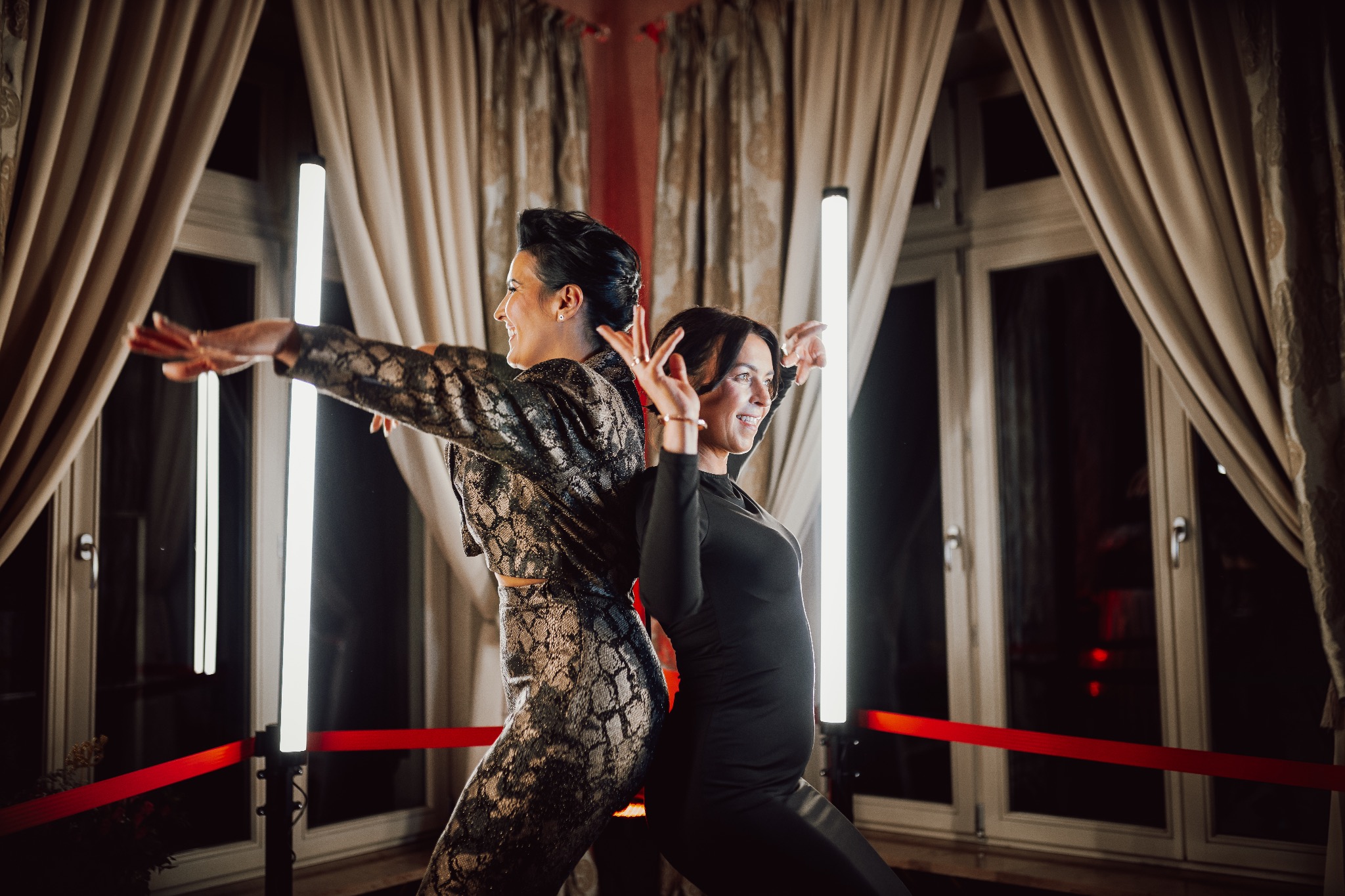 Dwie uśmiechnięte kobiety pozują tyłem do siebie na tle okien i lamp w eleganckim wnętrzu, sugerującym sesję zdjęciową lub zabawę.