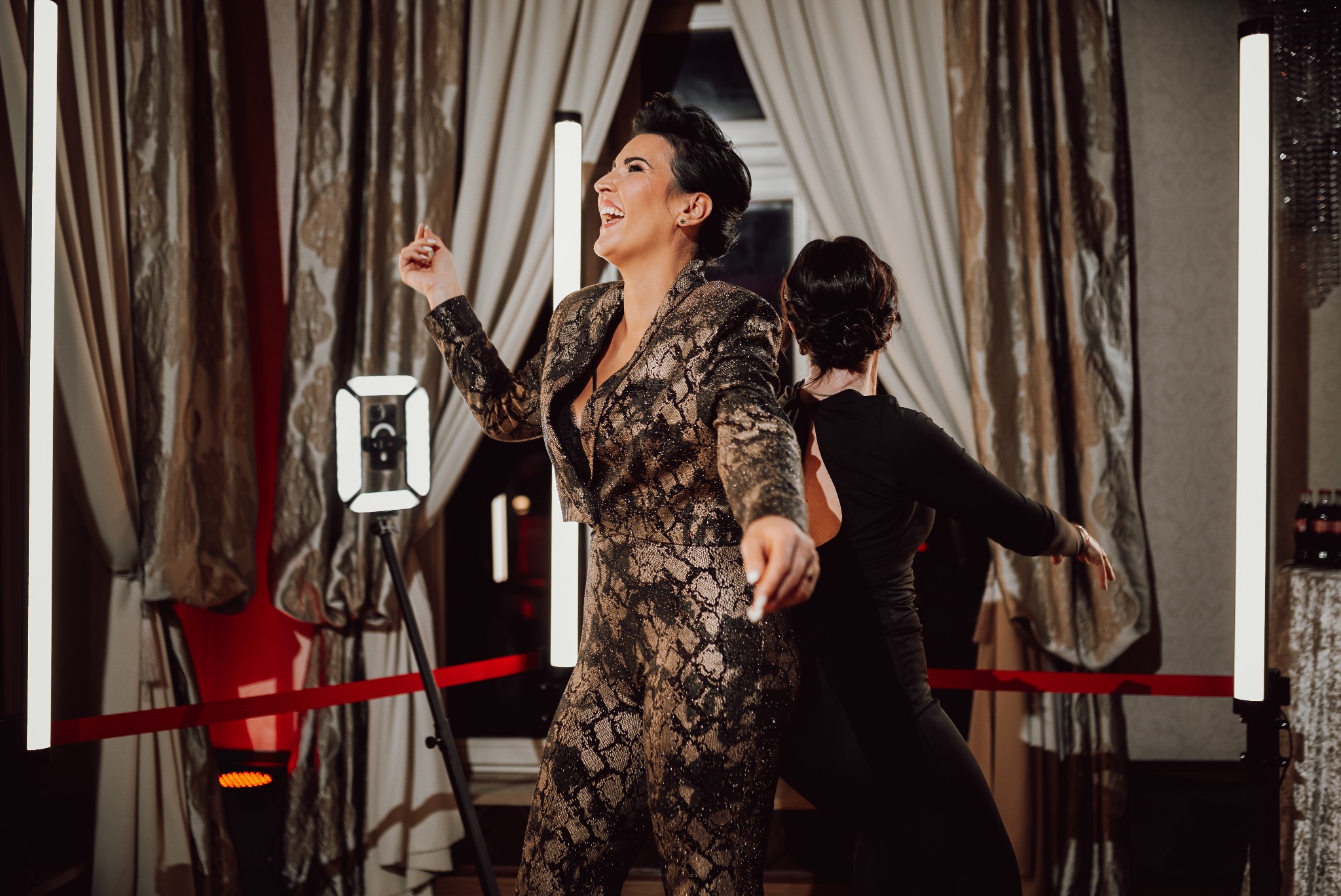 Rozbawiona kobieta w cekinowym kombinezonie tańczy na tle fotobudki z czerwonym sznurkiem i dekoracyjnymi zasłonami, druga osoba w tle.