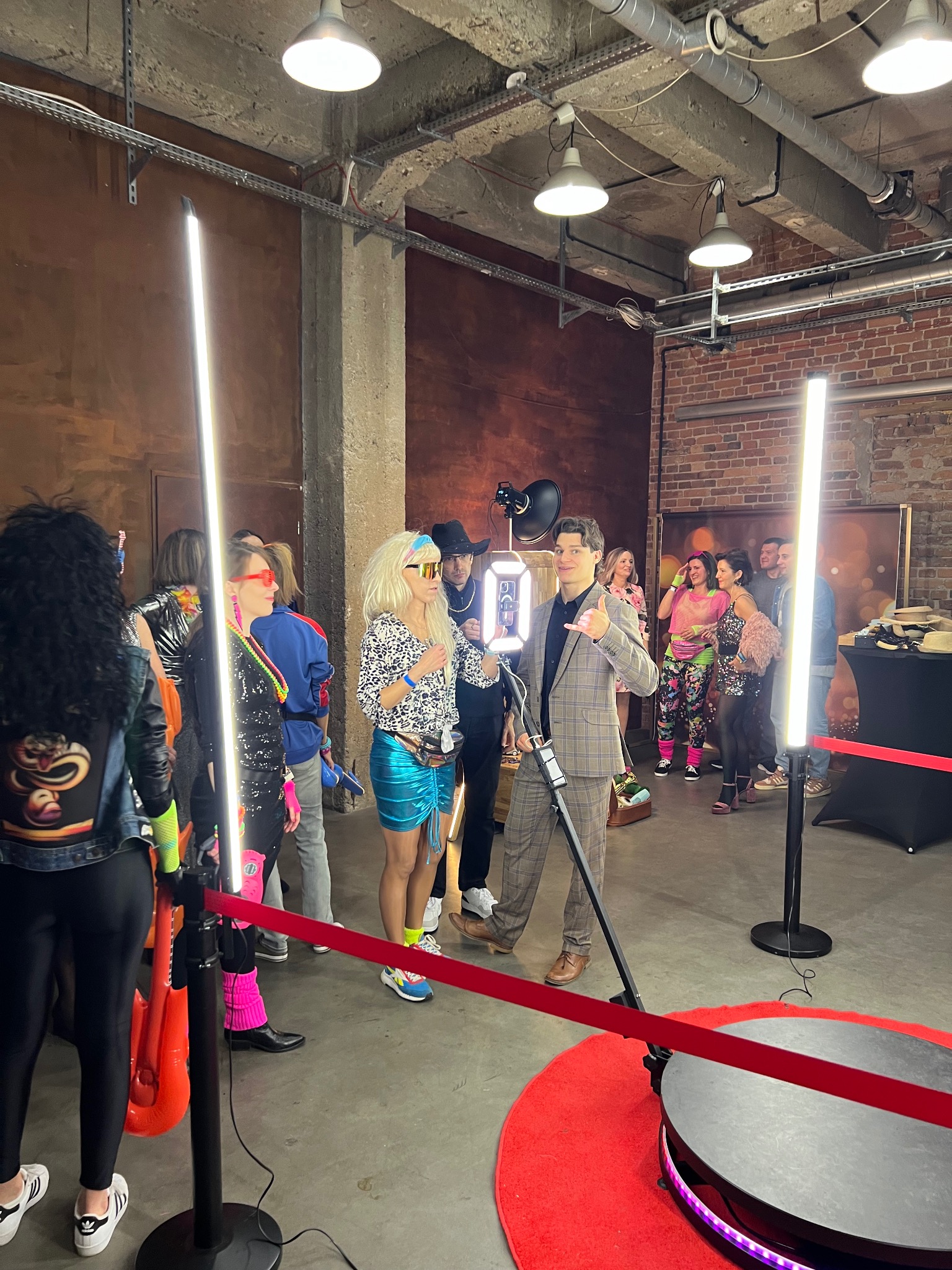 Uczestnicy w przebraniach z lat 80. pozują przy obrotowej platformie do zdjęć na tle ceglanej ściany w industrialnym wnętrzu. Oświetlenie ledowe.