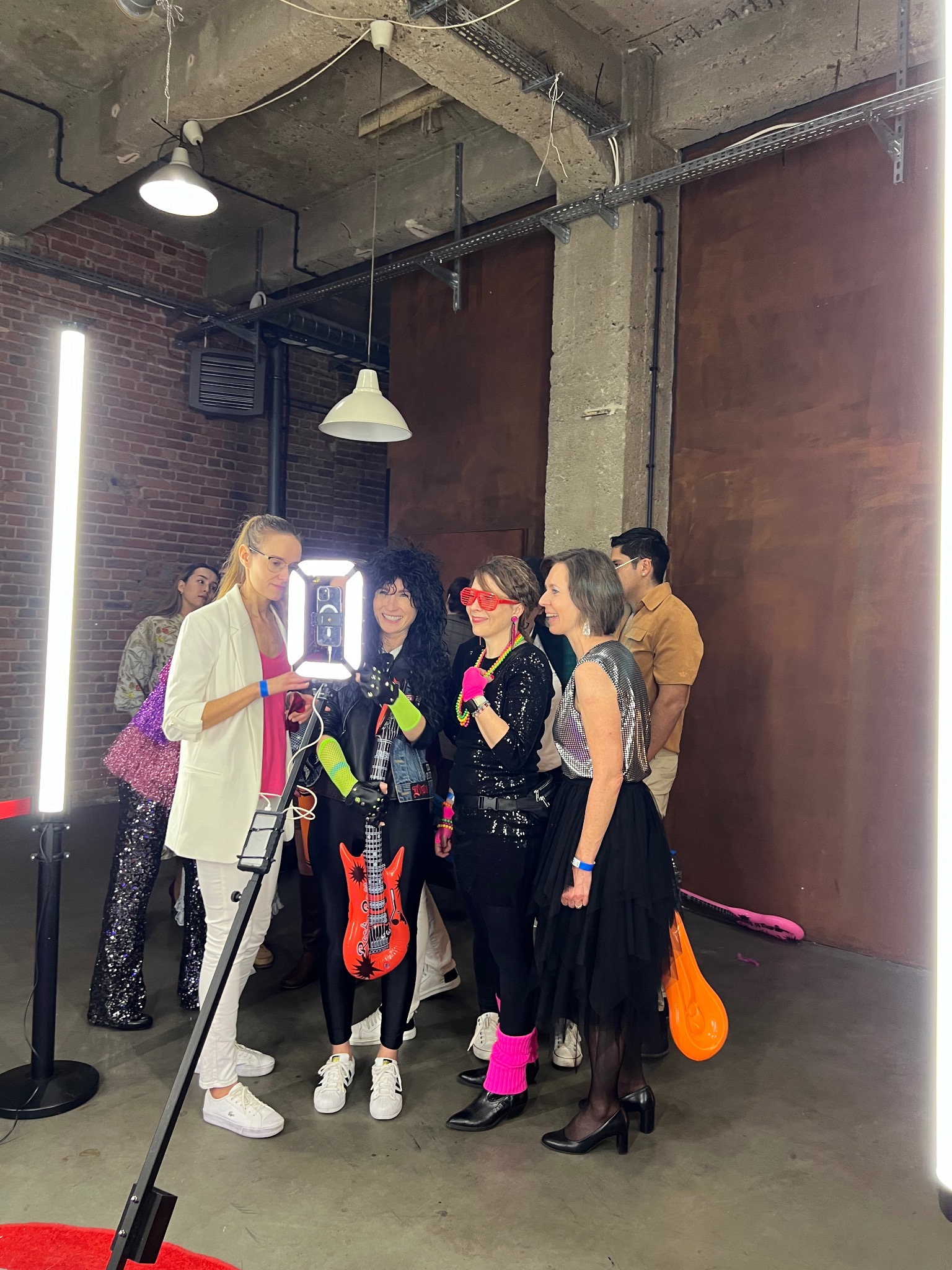 Grupa osób, w stylizowanych strojach, pozuje do zdjęcia przy stanowisku z oświetleniem LED i uchwytem na telefon, w loftowym wnętrzu.