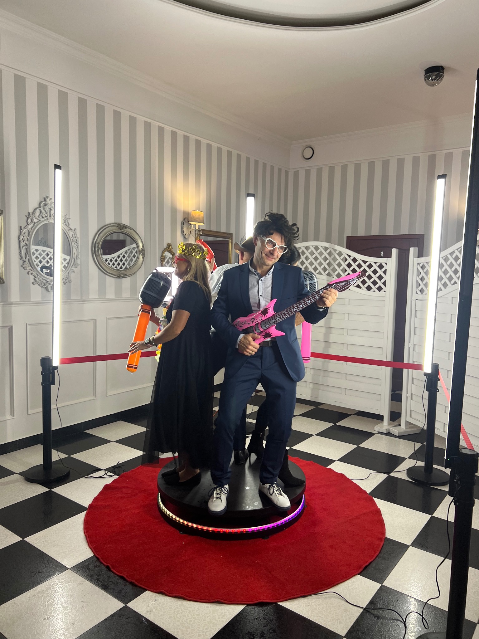 Dwie osoby pozują z rekwizytami (mikrofon i gitara) na platformie 360 fotobudki podczas imprezy. W tle ściana w paski i dekoracyjne lustra.