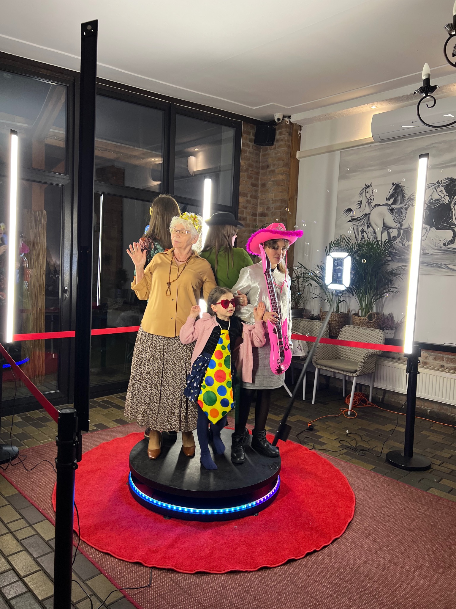 Radosna grupa osób w przebraniach pozuje w fotobudce z oświetleniem LED i rekwizytami, tworząc zabawną pamiątkę z imprezy.