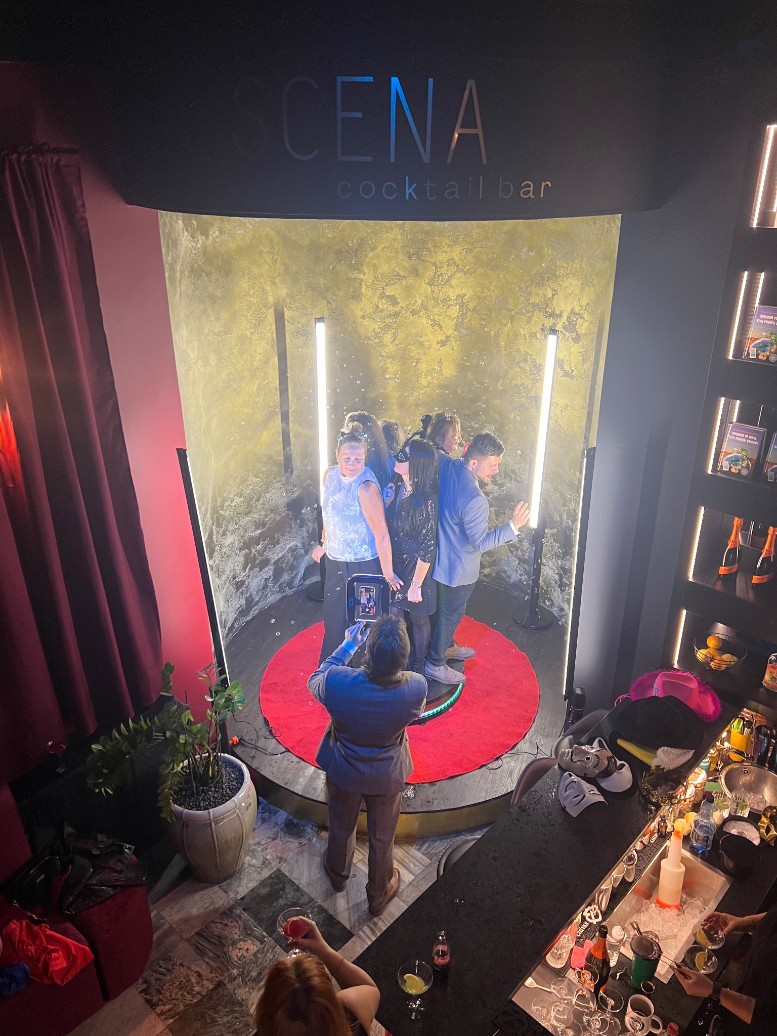 Grupa osób pozuje w fotobudce w barze 'Scena Cocktail Bar', fotograf robi zdjęcie telefonem. Widok z góry, w tle bar z akcesoriami i napojami.