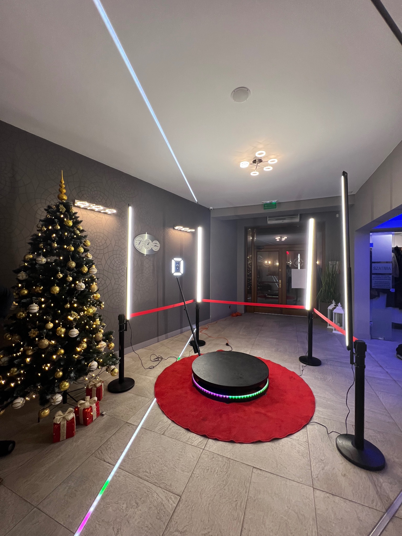 Okrągła platforma obrotowa na czerwonym dywanie, otoczona lampami LED i czerwoną taśmą, w tle choinka i zegar. Dekoracje świąteczne i oświetlenie.
