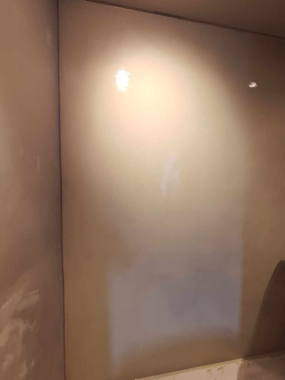 Gładka, beżowa ściana w rogu pomieszczenia, oświetlona punktowo, z widocznymi refleksami światła.