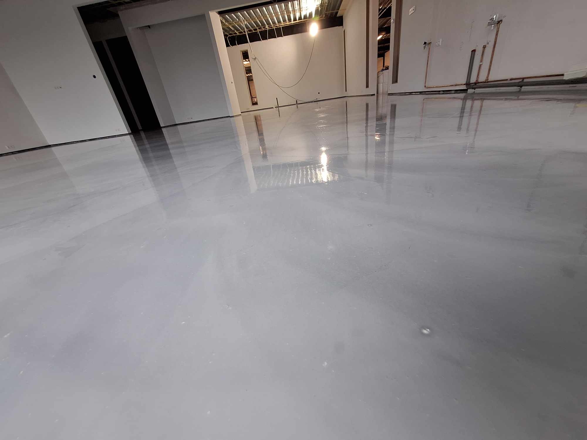 Błyszcząca, świeżo wylana szara posadzka żywiczna w surowym wnętrzu, widoczne rury instalacji na ścianie i niedokończony sufit.