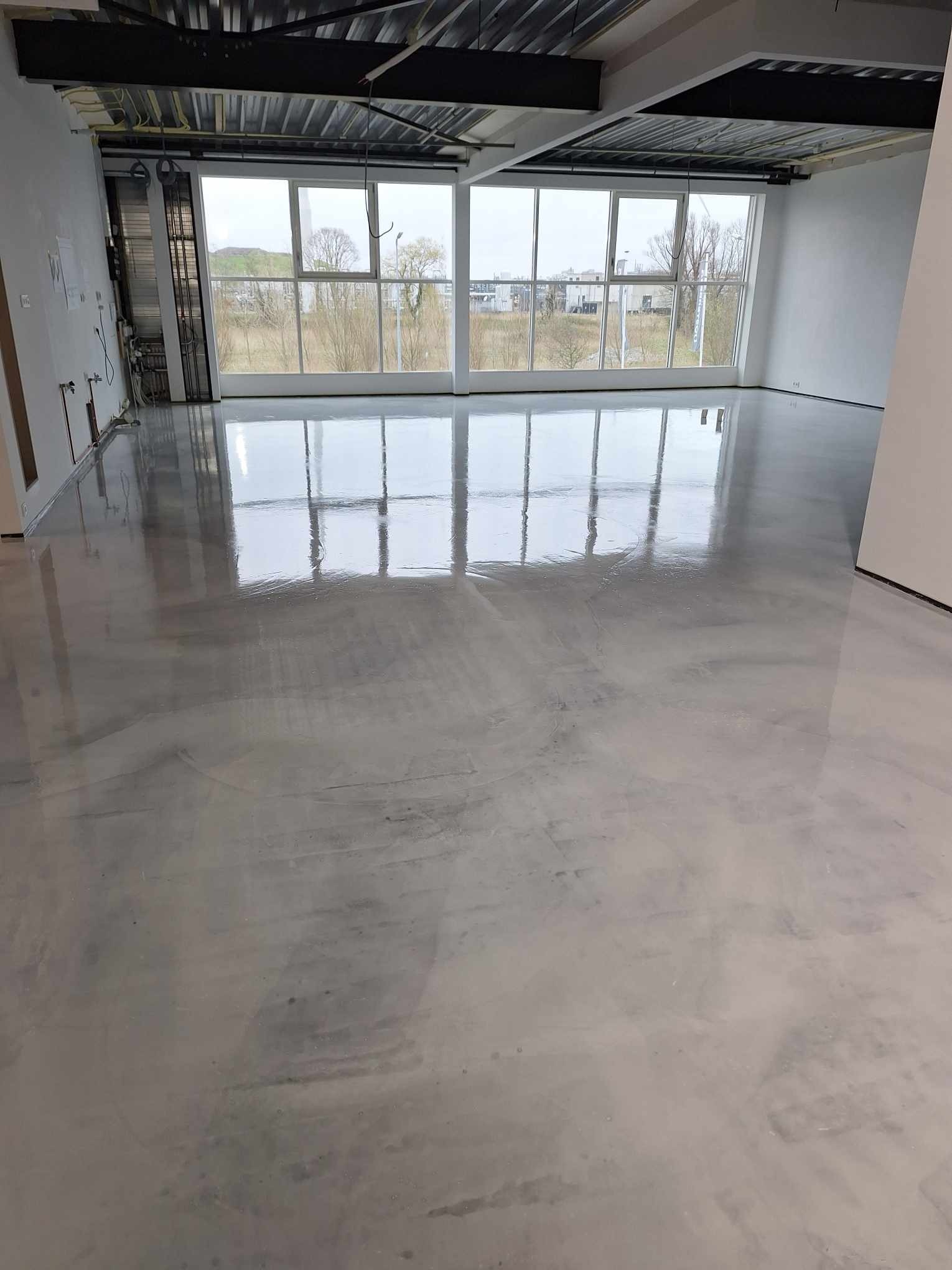 Wnętrze z nową, błyszczącą, szarą posadzką wylewaną, odbijającą światło z dużych okien. Widoczne niedokończone ściany i instalacje pod sufitem.