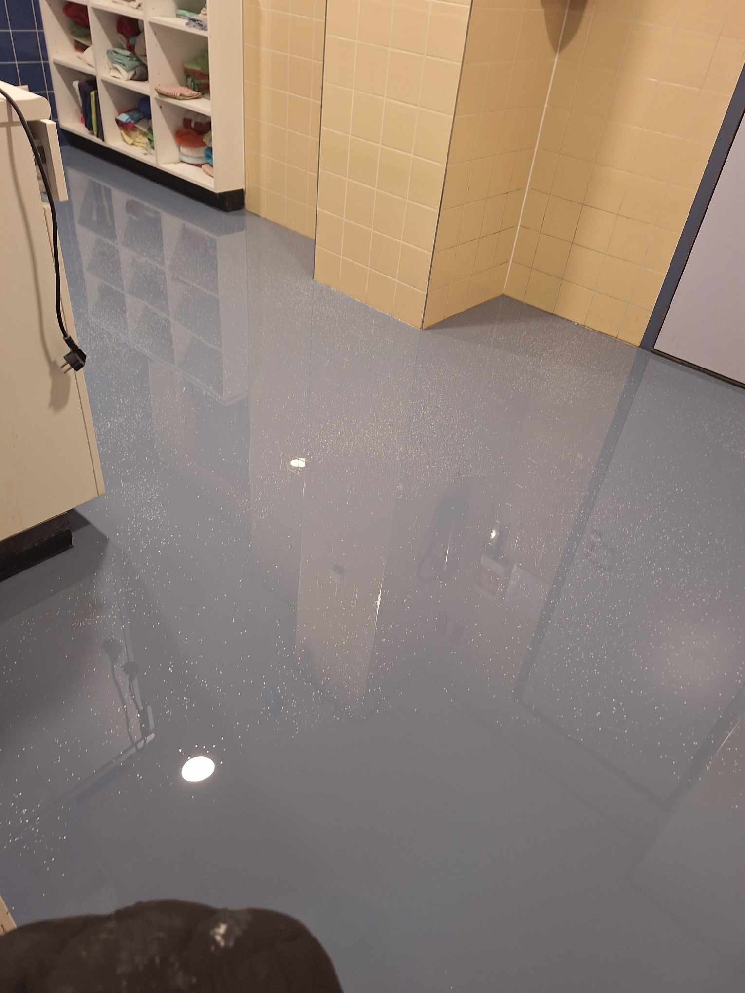 Błękitna, błyszcząca posadzka z delikatnym wzorem, widoczna w pomieszczeniu z jasnymi, kafelkowymi ścianami i białymi regałami.