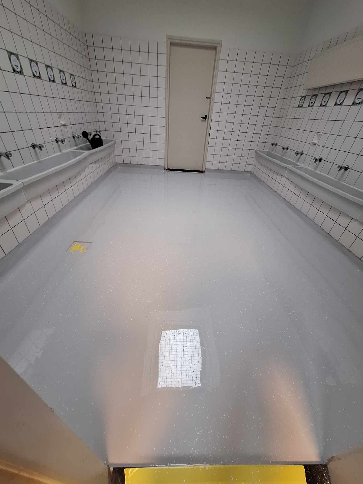 Szara, błyszcząca posadzka w pomieszczeniu z białymi, kwadratowymi płytkami na ścianach i rzędami umywalek. Widoczne odbicie okna w posadzce oraz żółte zabezpieczenie przy podłodze.