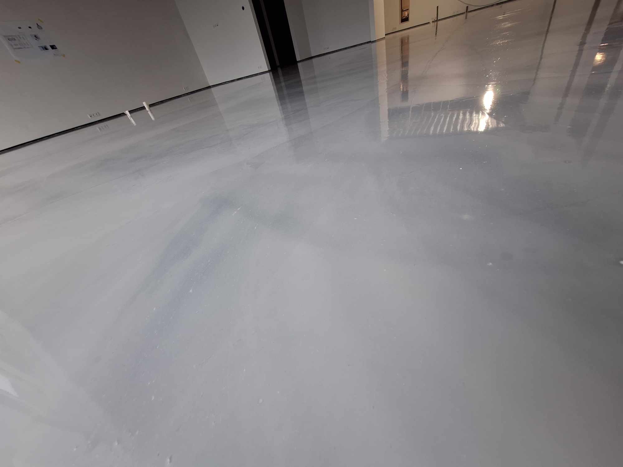 Świeżo wylana, gładka, szara posadzka z połyskiem w pustym pomieszczeniu, widoczne odbicia światła i elementów otoczenia.